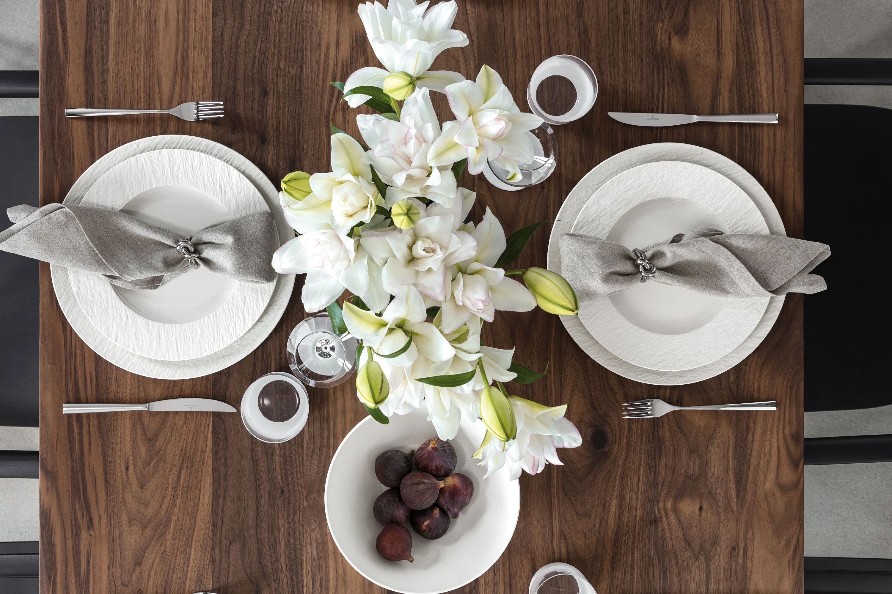 Vogelperspektive auf einen elegant gedeckten Walnussholztisch mit strukturierten weißen Manufacture Rock Blanc Porzellantellern, grauen Leinenservietten mit silbernen Ringen, einem natürlichen Centerpiece aus weißen Lilien und einer weißen Schale voller frischer, dunkler Feigen.