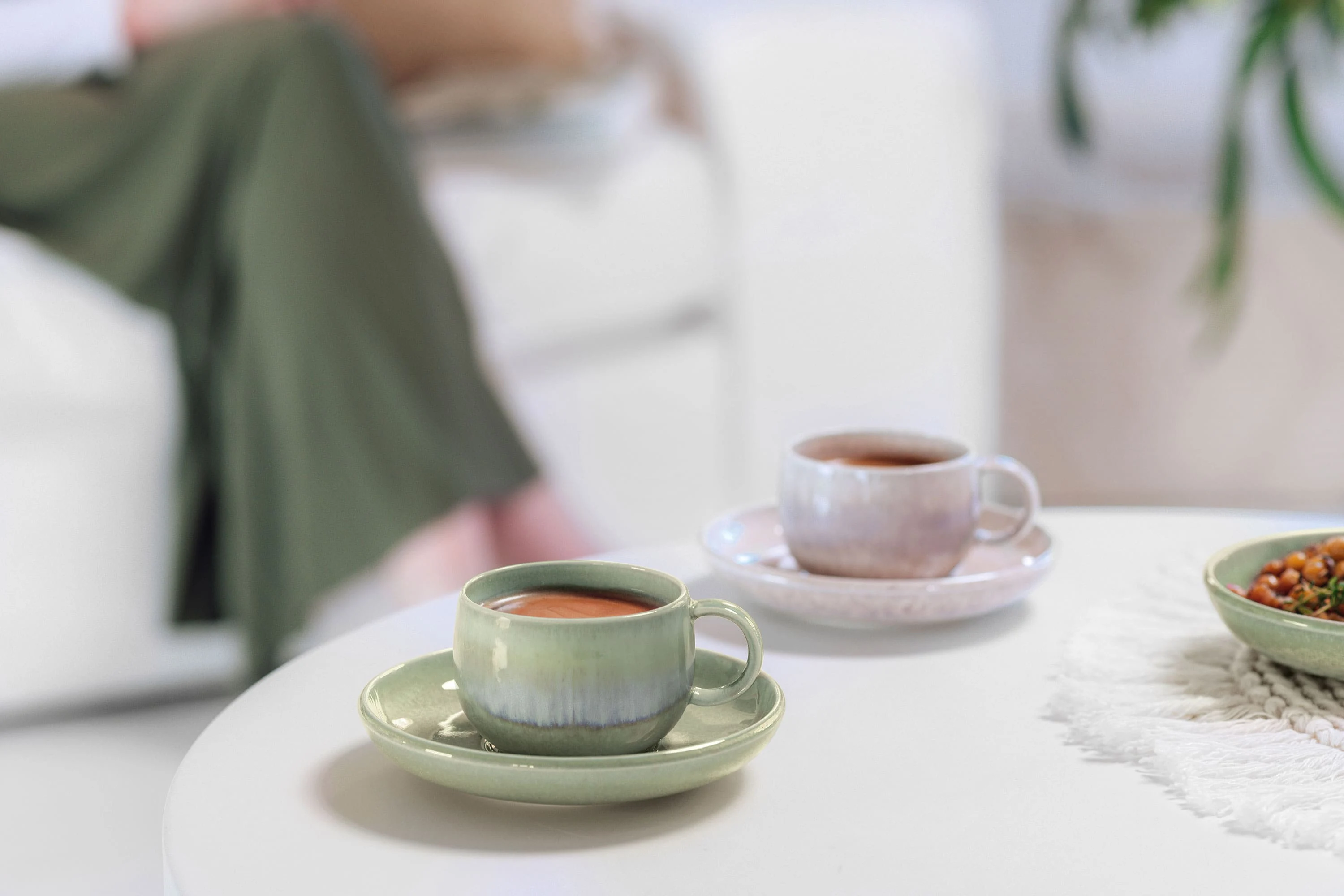 Two Perlemor coffee cups on a living room table, with a person partially visible on the sofa in the background.