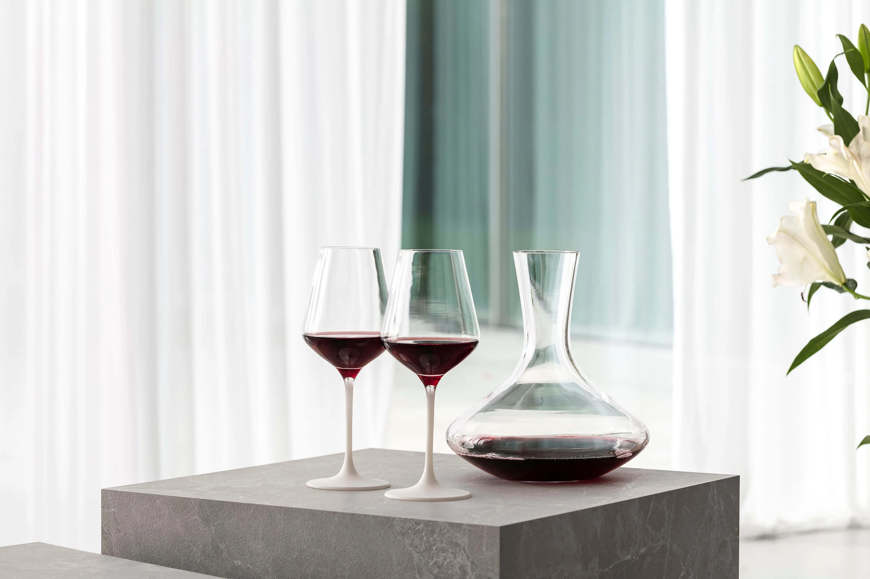 Two filled Manufacture red wine glasses on a table next to an elegant Purismo wine decanter.