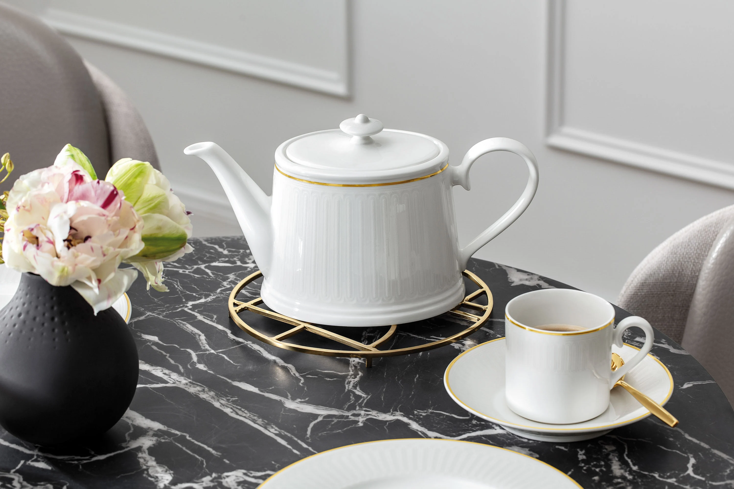 A white coffee pot with a gold rim on a stand on a marble table. Next to it is a matching teacup and saucer with a gold spoon and a black vase with a bouquet of flowers.
