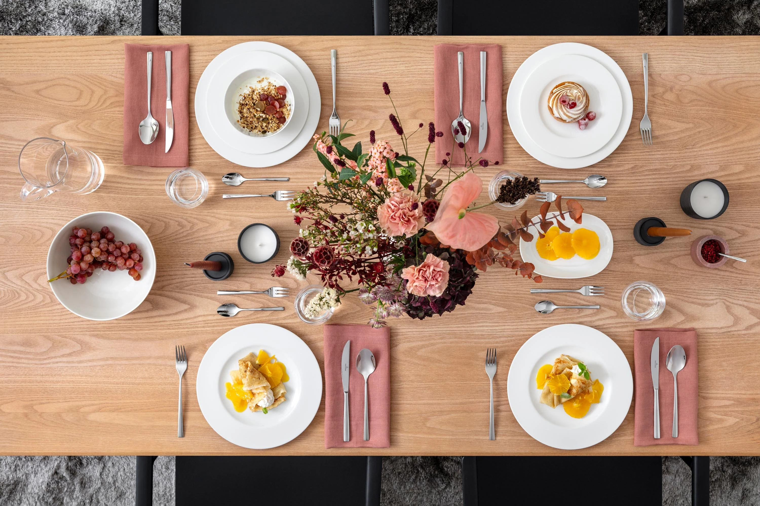 Florales Tablesetting mit Royal von Villeroy & Boch Ein rechteckiger Holztisch ist für vier Personen gedeckt. Es stehen Teller mit Essen, rosa Servietten, Besteck, Kerzen und ein Blumengesteck auf dem Tisch. Trauben und Gewürze stehen ebenfalls auf dem Tisch.