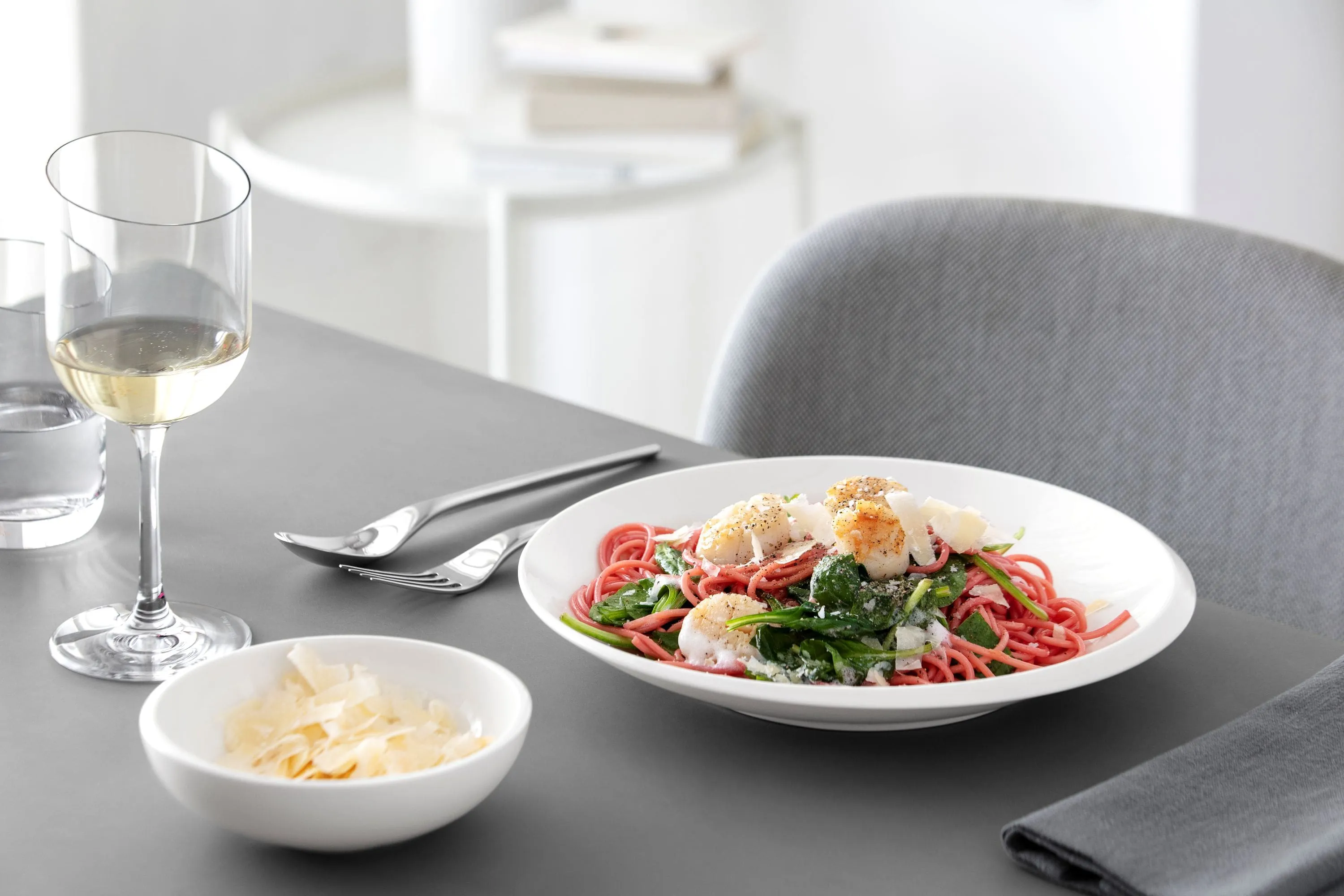 A plate of pasta, vegetables, cheese, a bowl of grated cheese, and a glass of white wine on a gray table.