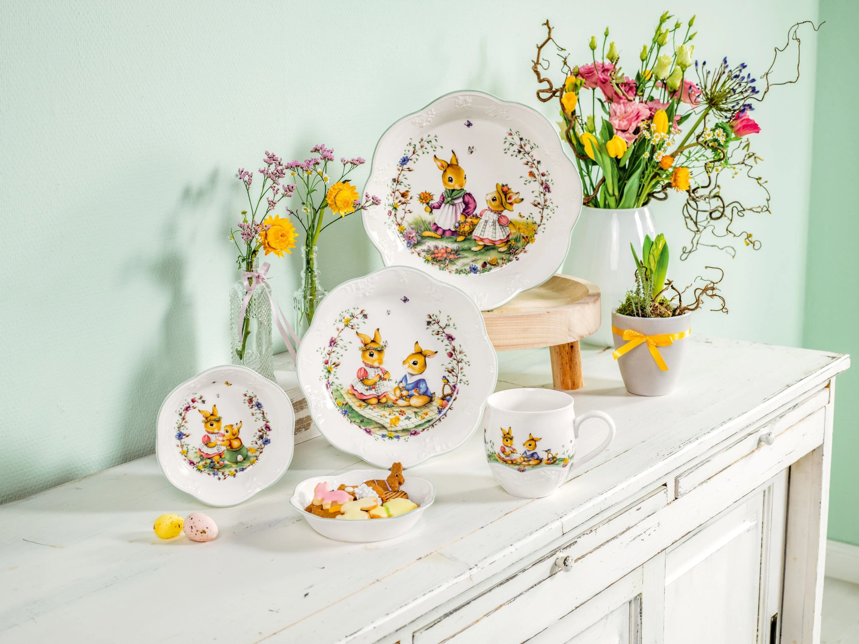 Four easter bowls decorated with bunnies, an easter mug and a some flower pots with spring flowers in them all sitting on a white wood dining sideboard