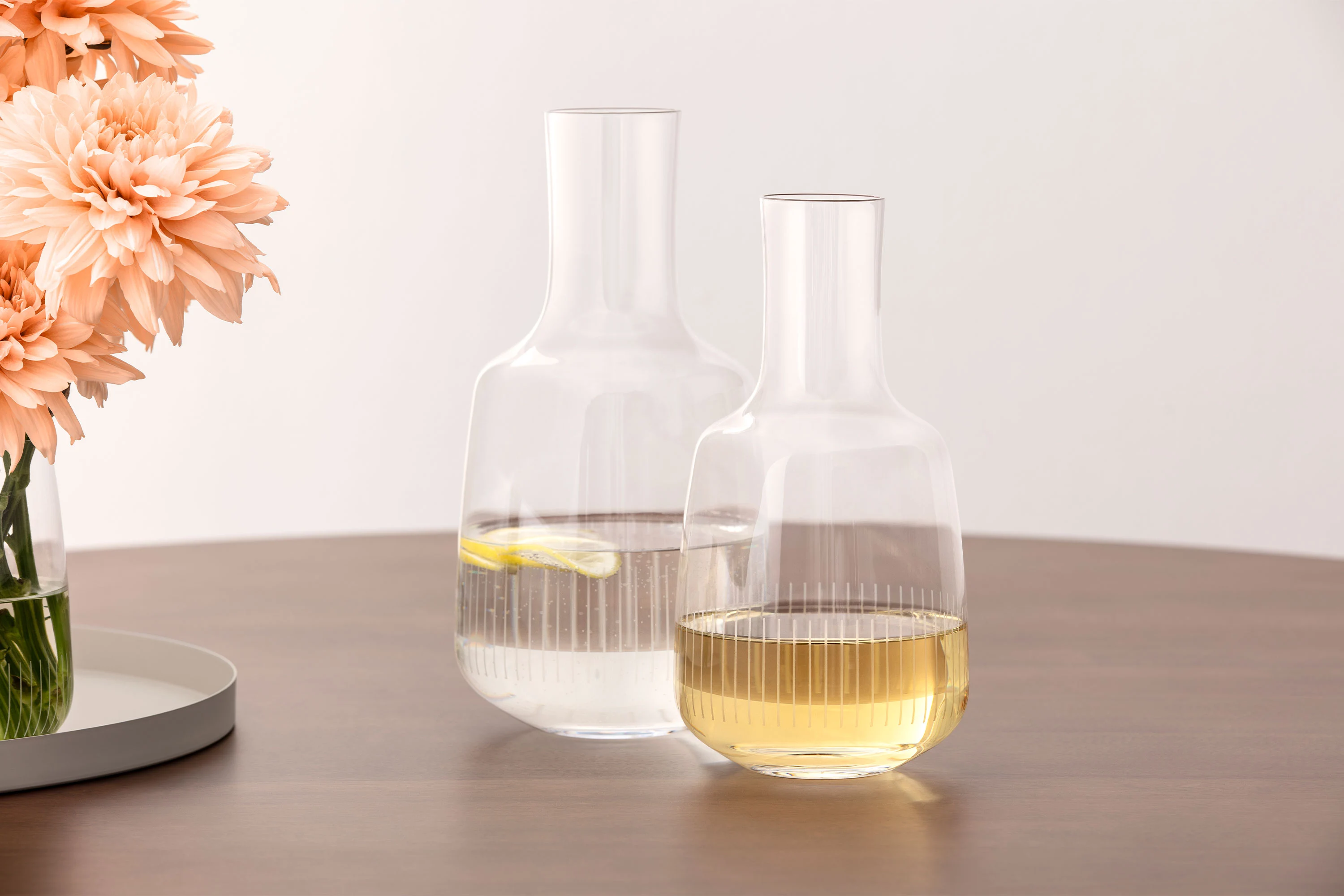Two glass carafes, one with water and a slice of lemon, one with a pale yellow liquid, on a wooden table next to flowers.
