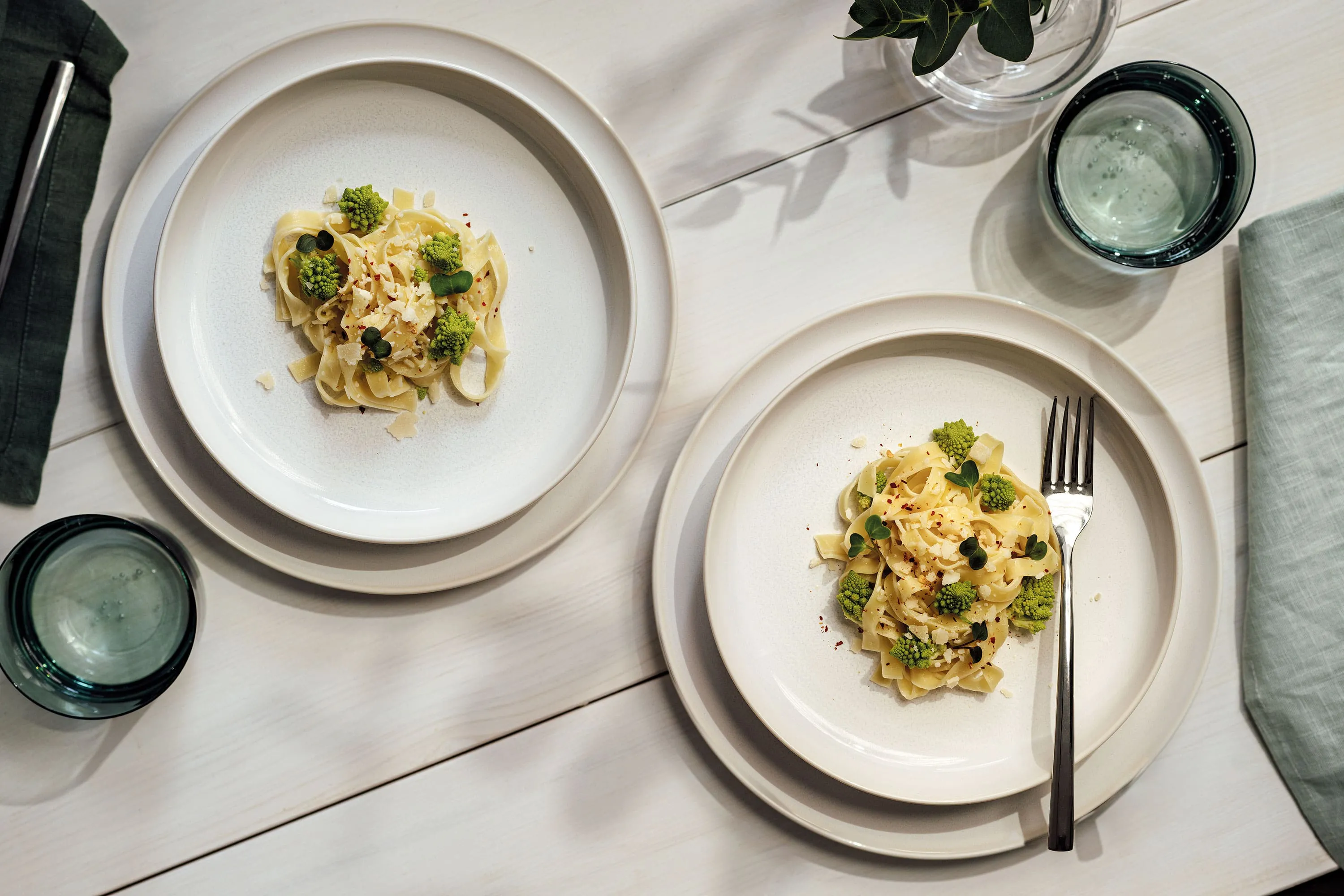 Two Crafted plates with broccoli and sauce on a white table, with glasses of water and a small potted plant.