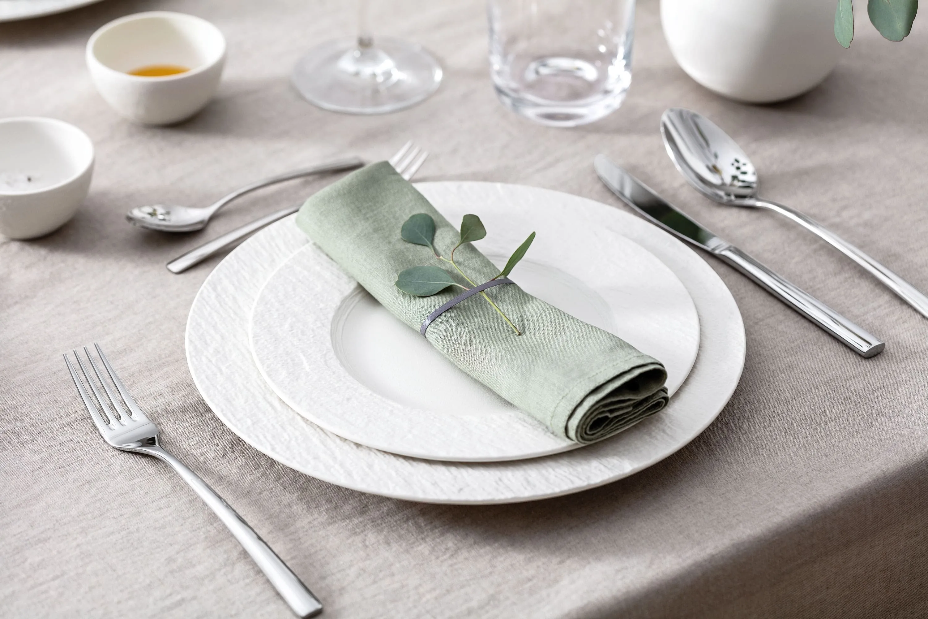 Des assiettes blanches Manufacture avec une serviettes sur une table Table dressée avec deux assiettes blanches Manufacture, une serviette vert pastel, des couverts de table Piemont et de l’eucalyptus.