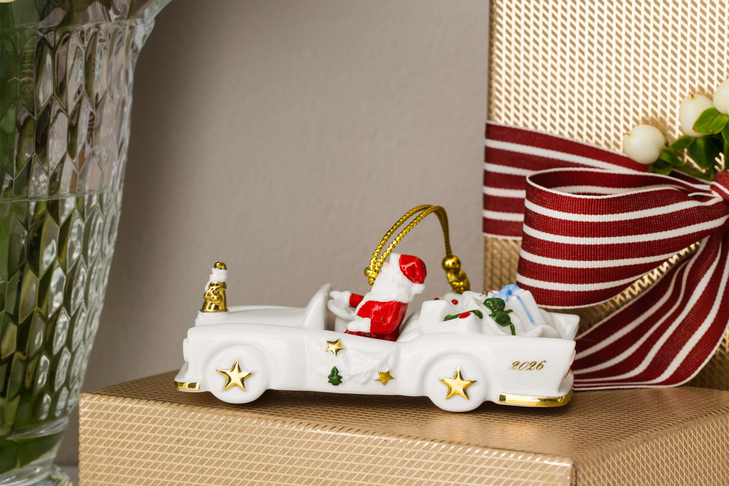 A ceramic Christmas car with presents is sitting on a gift wrapped in gold, next to a vase of red and white flowers.