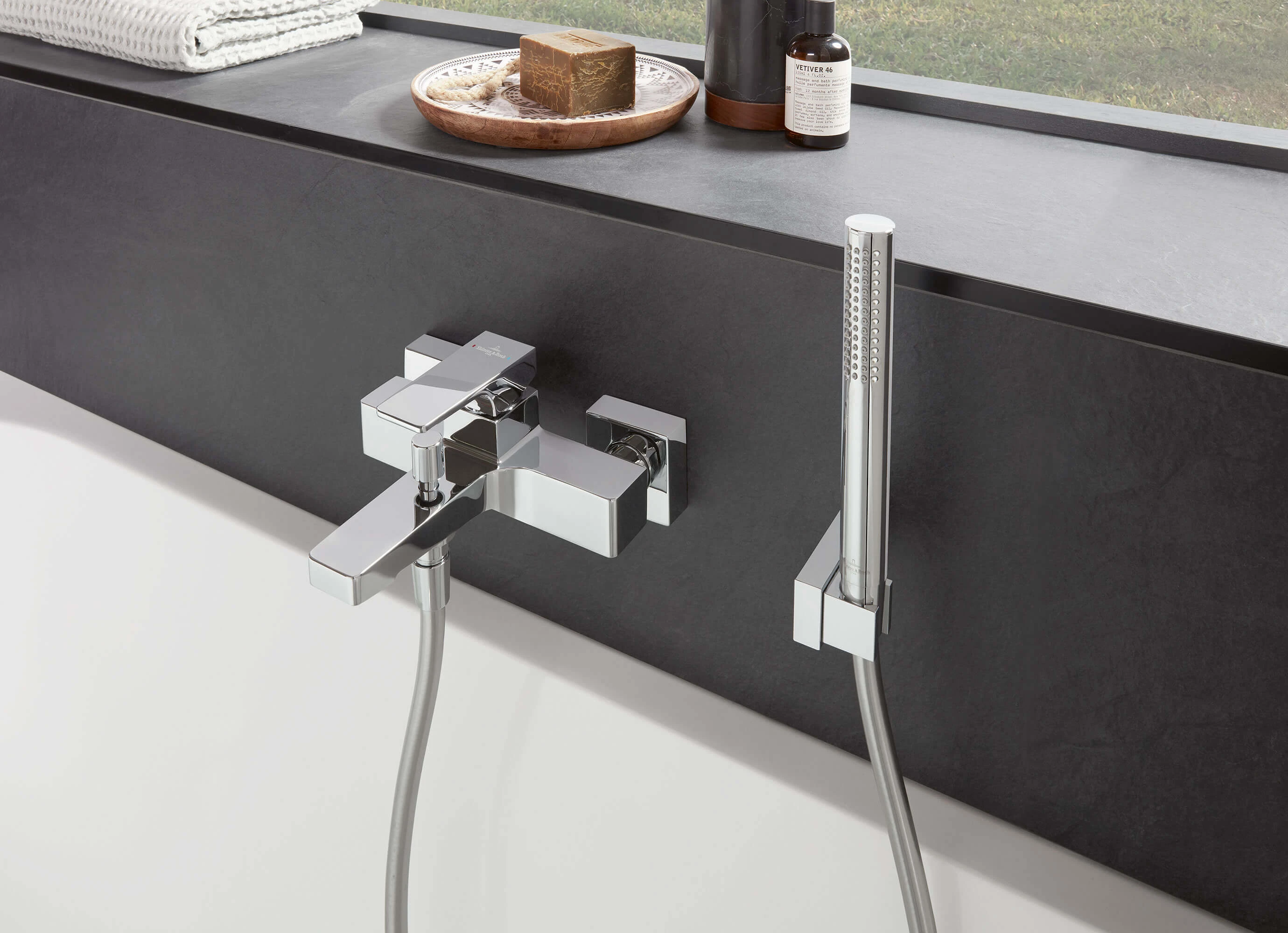 Architectura Square badekartermostat med dusjhode fra Verve Showers Nærbilde av en moderne badekartermostat med dusjhode foran en svart overflate. På bildet ser du et såpestykke, et hvitt håndkle og en flaske på avsatsen bak kranen.