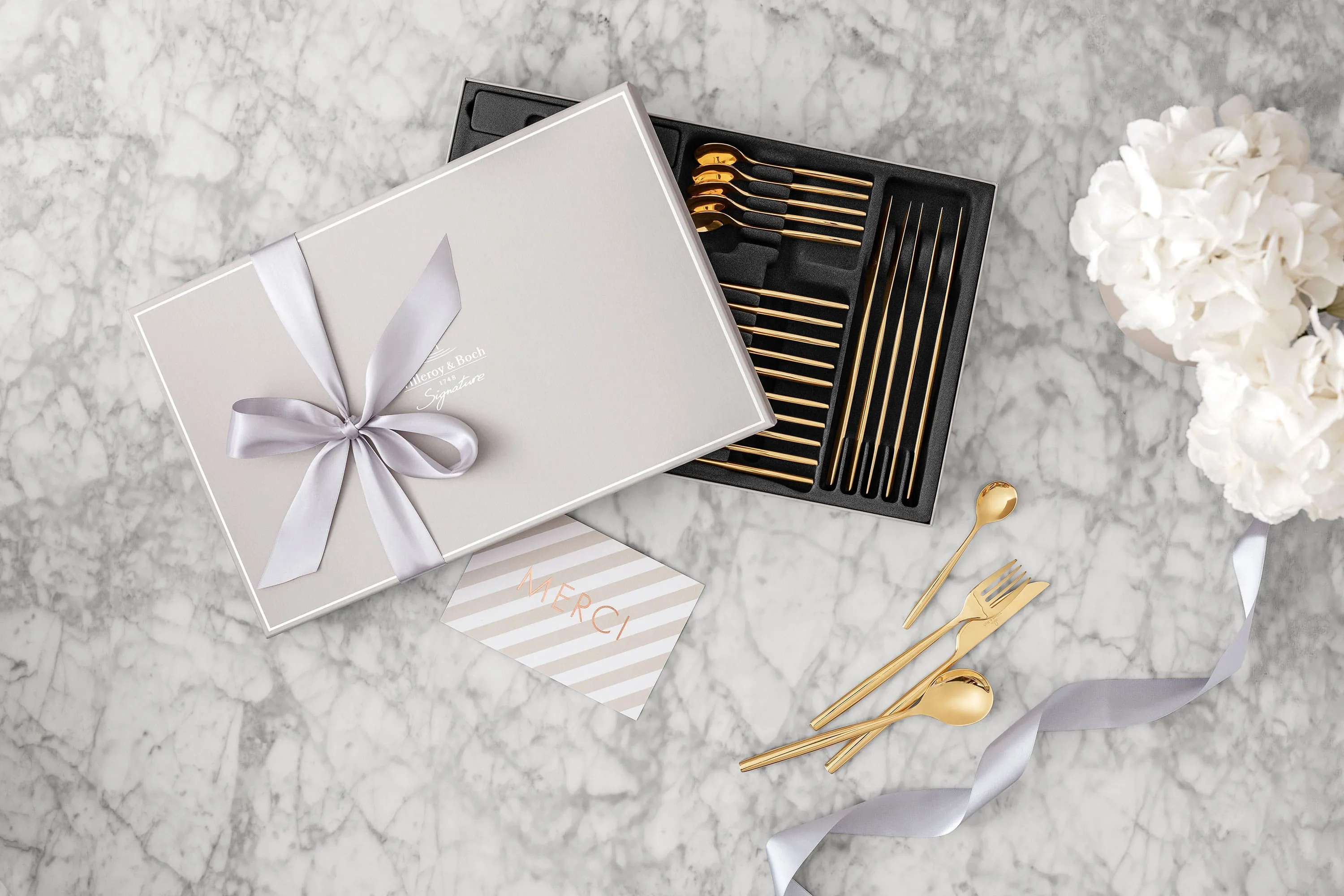 Une boîte cadeau avec des couverts de table MetroChic dorés et un ruban Une boîte cadeau avec des couverts de table MetroChic dorés et un ruban, une carte de remerciement et des fleurs blanches sur une surface en marbre.