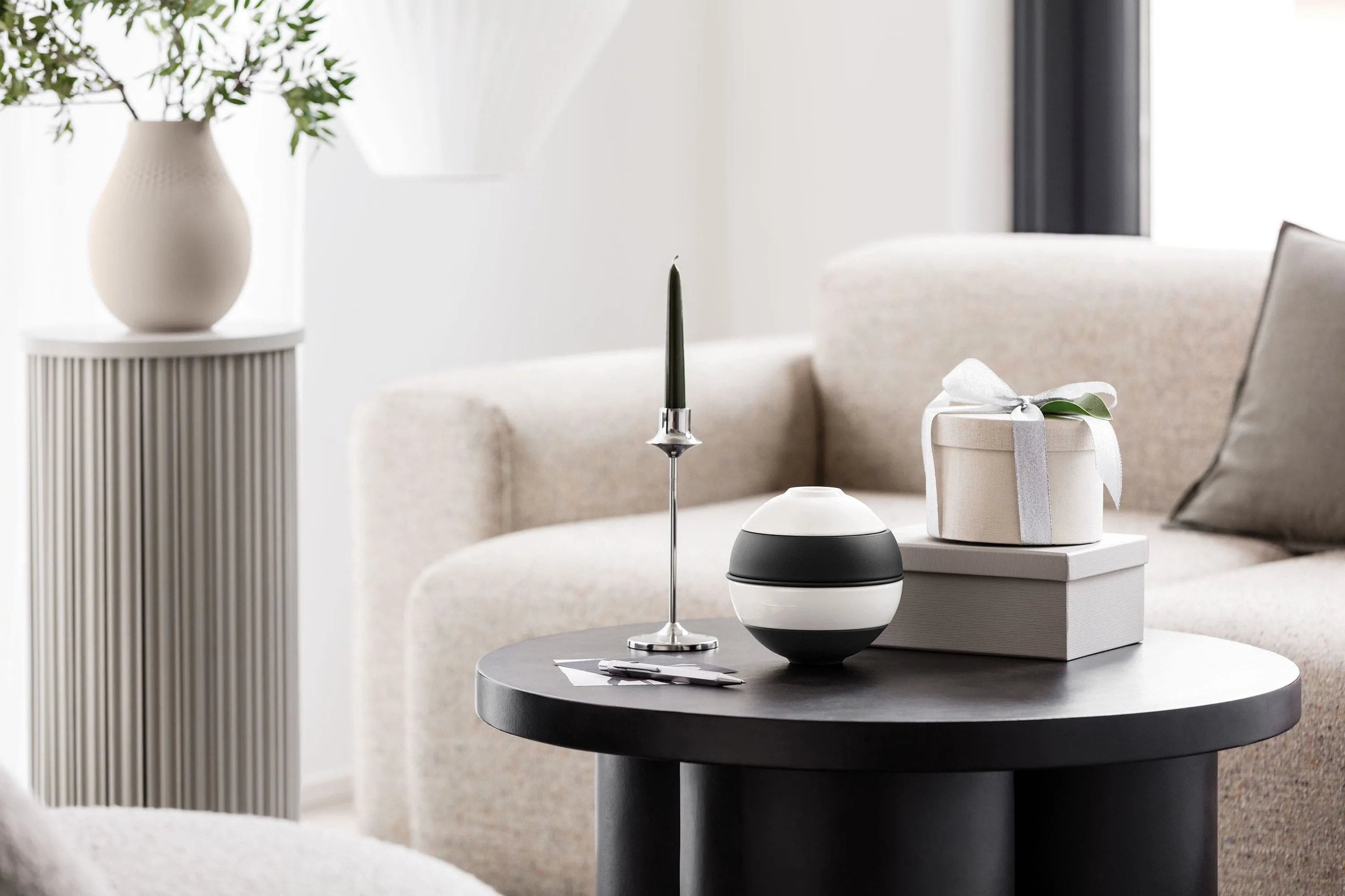 Une table basse ronde noire avec La petite Boule de Villeroy & Boch Une table basse ronde noire avec La petite Boule de Villeroy & Boch, une bougie verte sur un support argenté et une boîte cadeau sur deux livres. Un canapé beige et un socle gris avec un vase en arrière-plan.