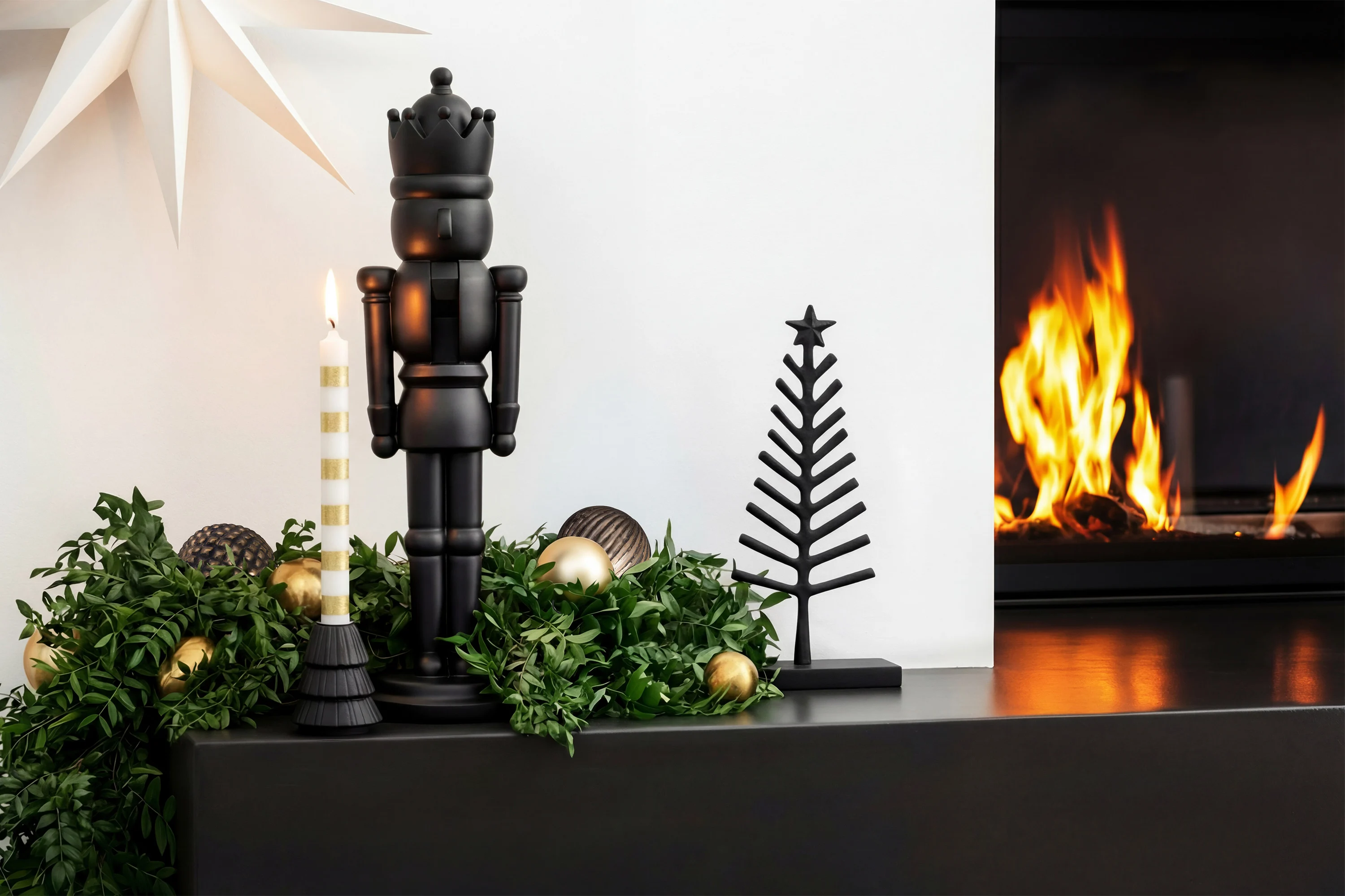 A black nutcracker, a candle and a metal tree A black nutcracker, a candle and a metal tree stand on a decorated mantelpiece next to a lit fire.