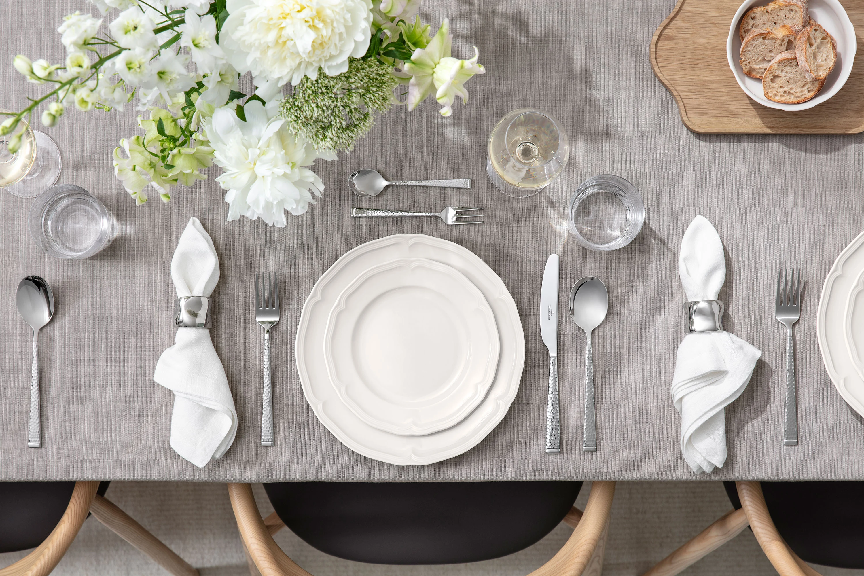 Der Tisch ist mit weißen Tellern, Servietten, Besteck, Gläsern und einem Blumenarrangement auf einem grauen Tischtuch gedeckt.