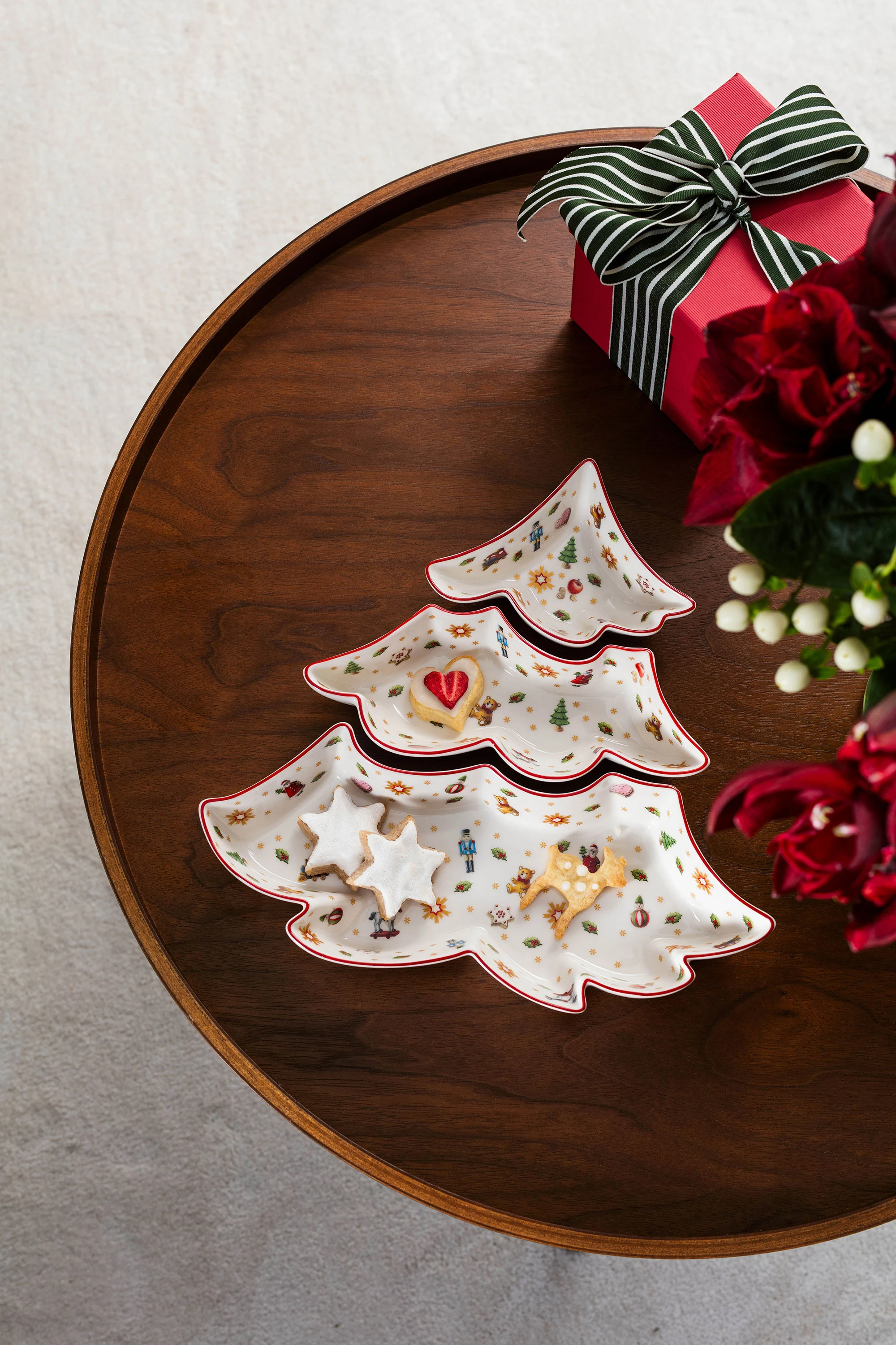 Overhead view of a 3-piece Christmas tree-shaped porcelain snack bowl set from Toy's Delight collection on a dark wooden table, filled with festive star cookies and a heart-shaped biscuit, next to a red gift box with a green-and-white striped ribbon and deep red flowers