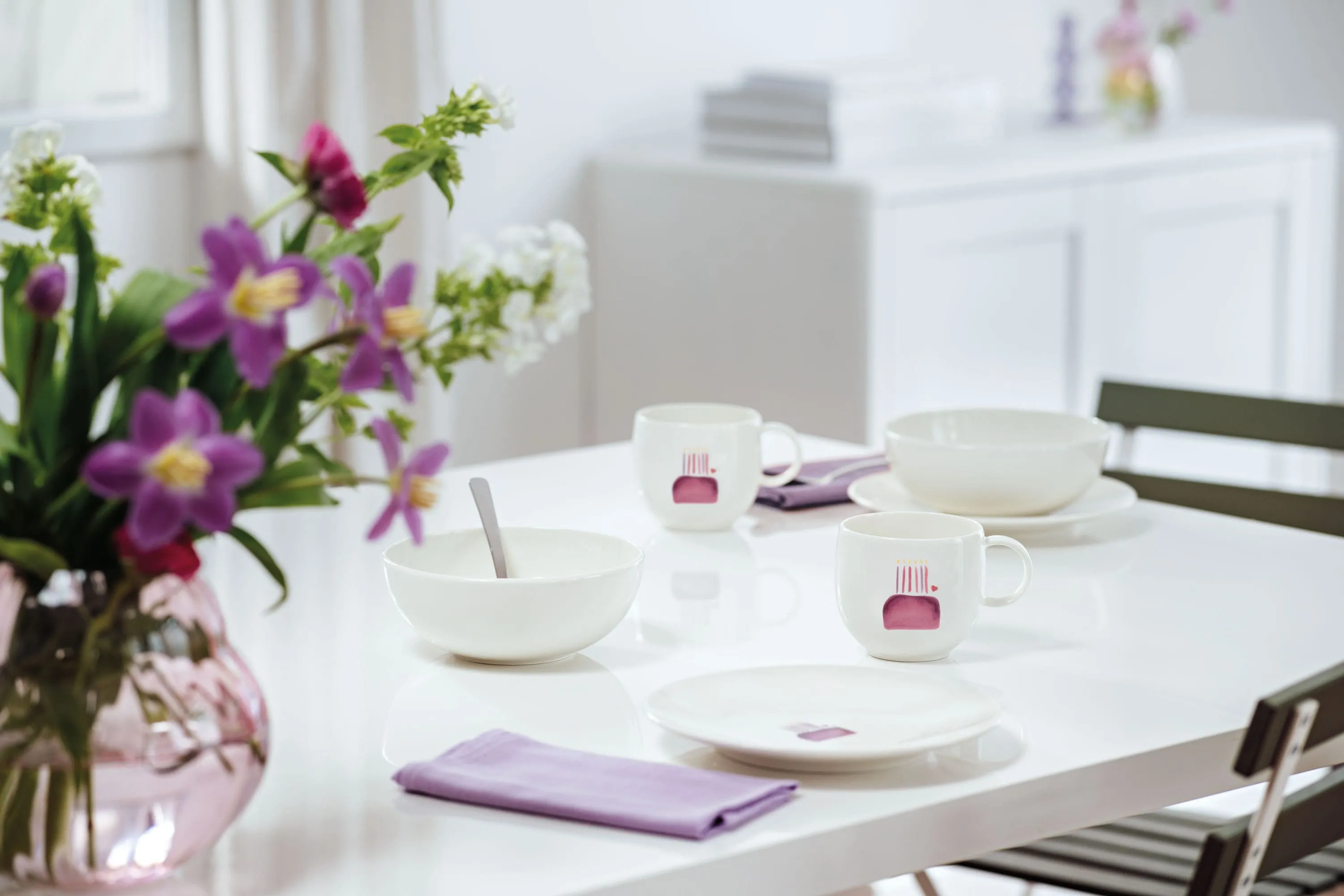 De la vaisselle With Love avec un motif d’anniversaire sur une table pour 2 personnes Une table blanche pour deux personnes avec des plats creux, des gobelets, des assiettes de la collection With Love avec un motif d’anniversaire et des serviettes de table couleur lavande. À gauche, se trouve un vase avec des fleurs violettes et blanches.