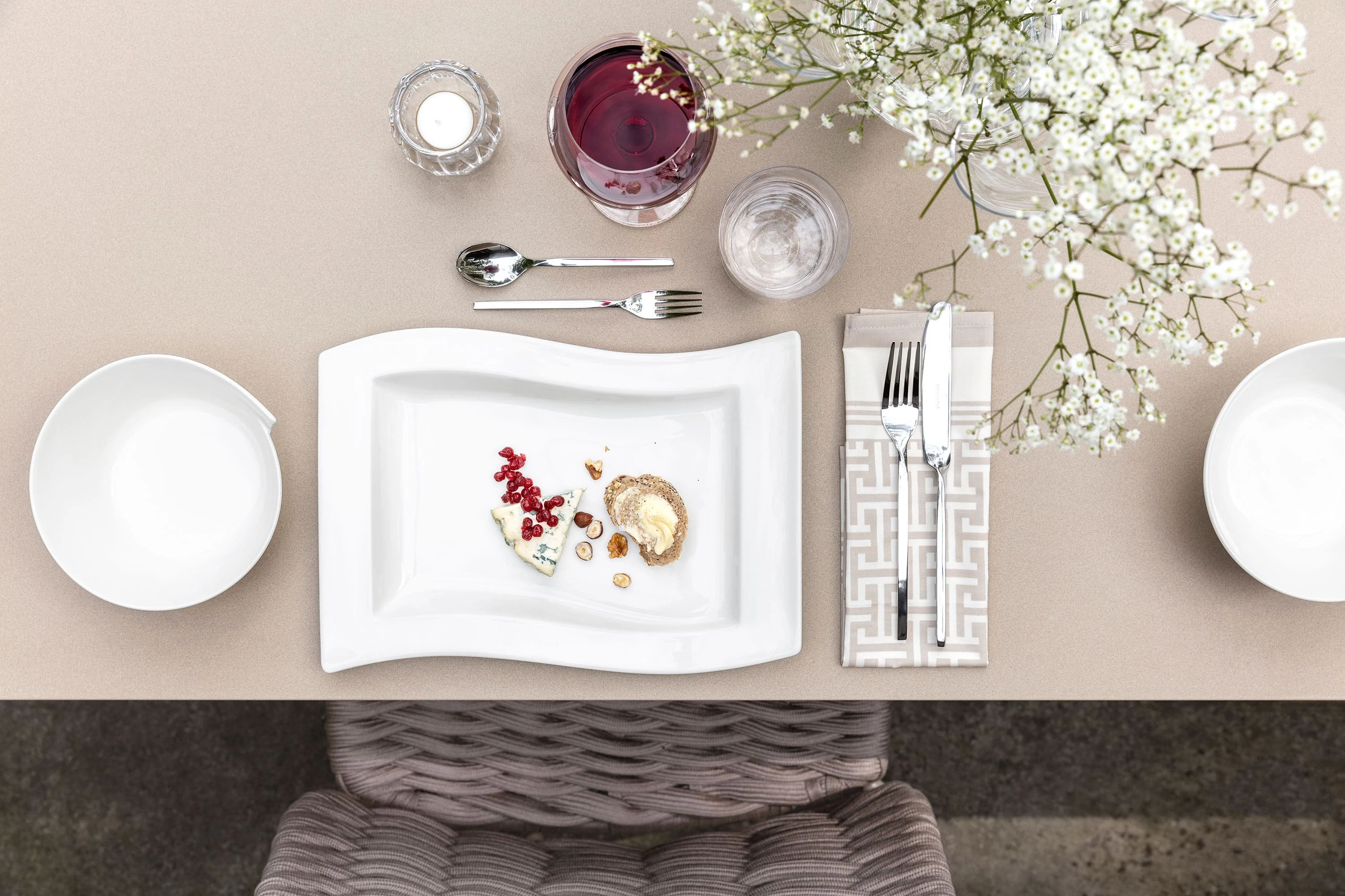 A sophisticated place setting with a modern white NewWave plate from Villeroy & Boch, which sets the scene for the food and is surrounded by cutlery, a wine glass and a water glass on a beige table. White flowers add an elegant touch to the scene.