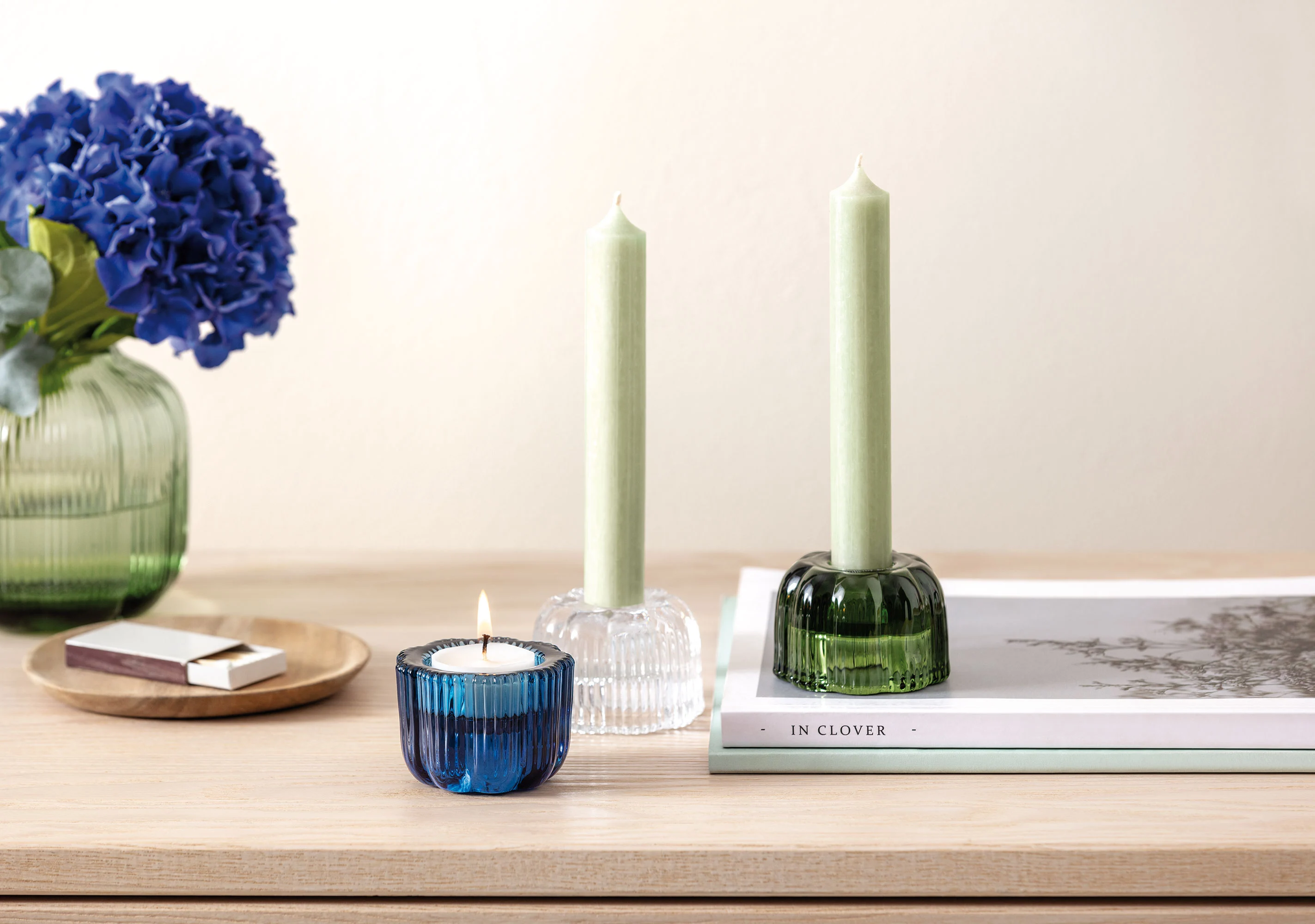 On a wooden table stands a Fleur vase and Fleur tealight holder from Villeroy & Boch a blue flower arrangement adorns the vase On a wooden table stands a Fleur vase and Fleur tealight holder from Villeroy & Boch, a blue flower arrangement adorns the vase, a burning candle in a blue holder, two green candles in holders, a book and matches.