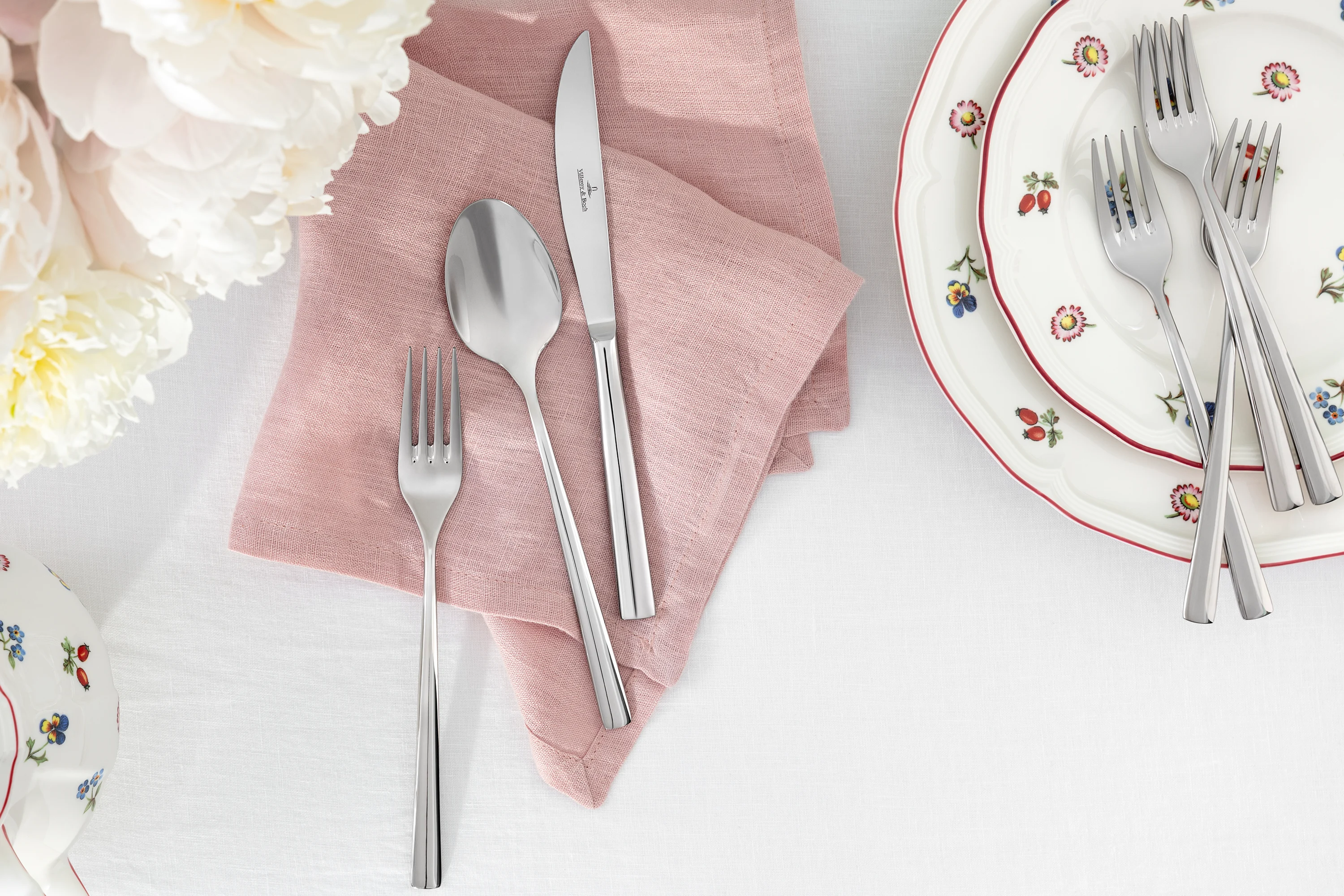 Piemont cutlery lies on a pink Lifestyle Accessories napkin next to stacked Petite Fleur plates on a white tablecloth.
