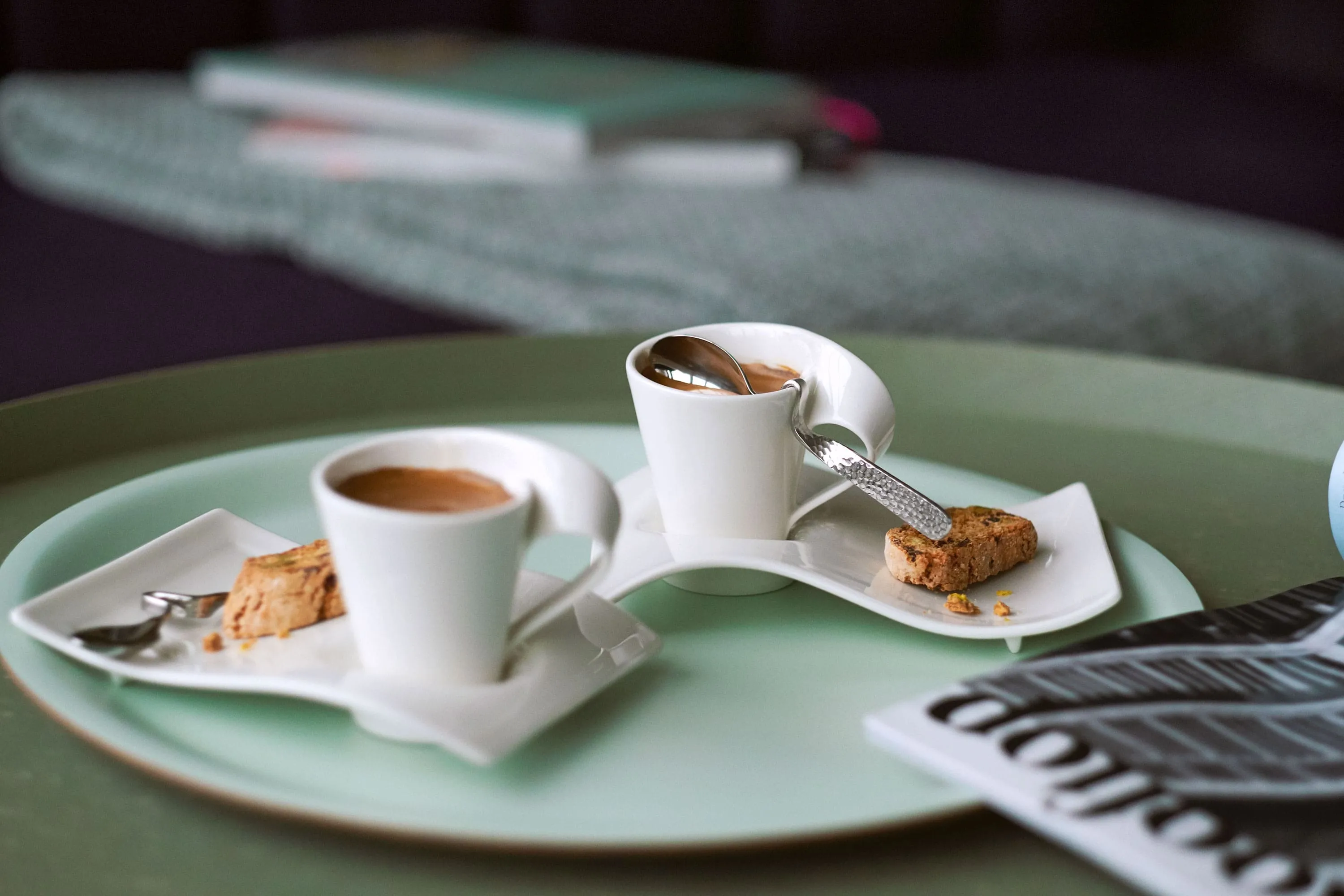 Deux tasses à expresso NewWave sur un plateau vert avec un journal Deux tasses à expresso NewWave avec des cuillère et des sous-tasses, sur un grand plateau vert avec un journal à côté.