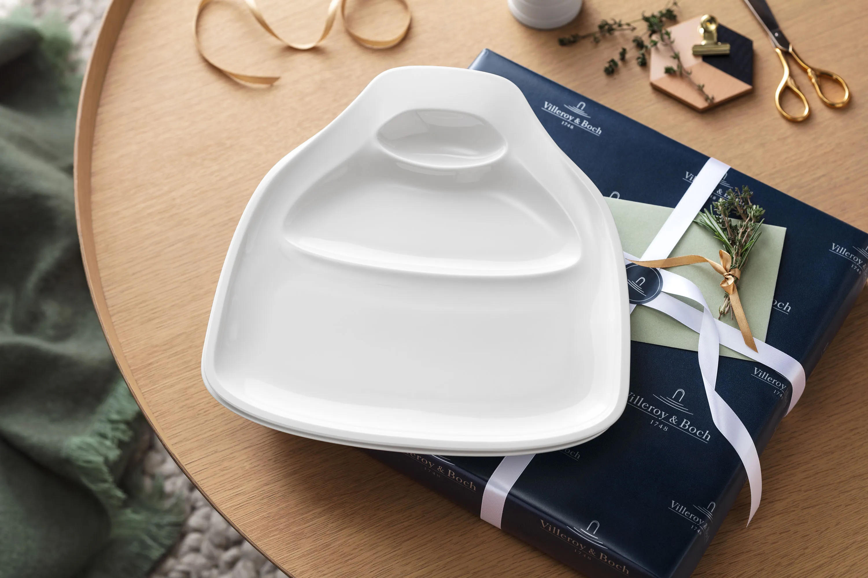 Une assiette BBQ Passion dans un emballage cadeau Une assiette BBQ Passion de Villeroy & Boch prête à offrir avec une boîte cadeau, un petit bouquet de romarin et de thym et des ciseaux dorés en arrière-plan.