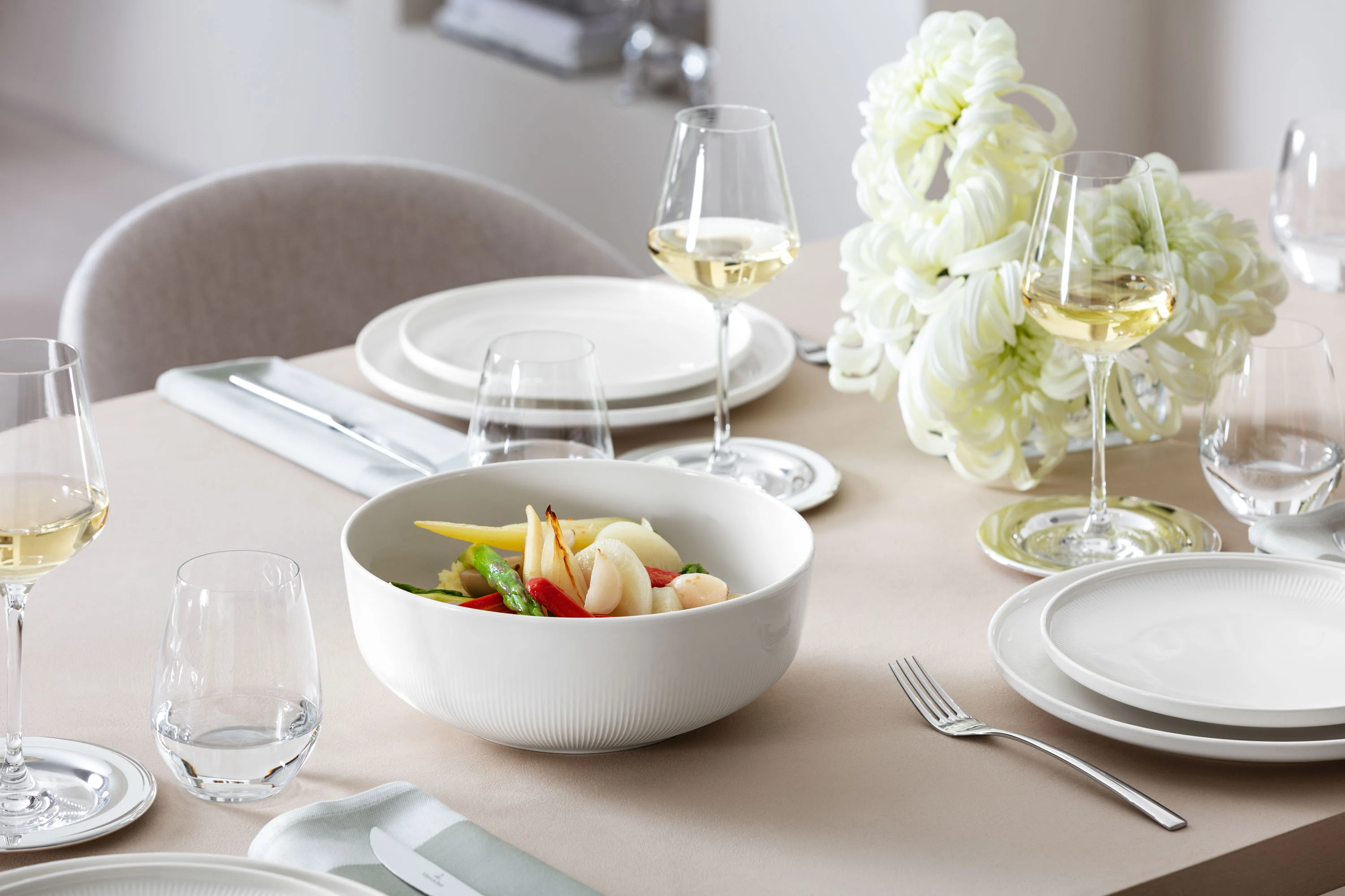White Afina bowl with vegetables on a dining table Large Villeroy & Boch Afina serving bowl filled with colourful vegetables on a laid table, with flowers in the background.