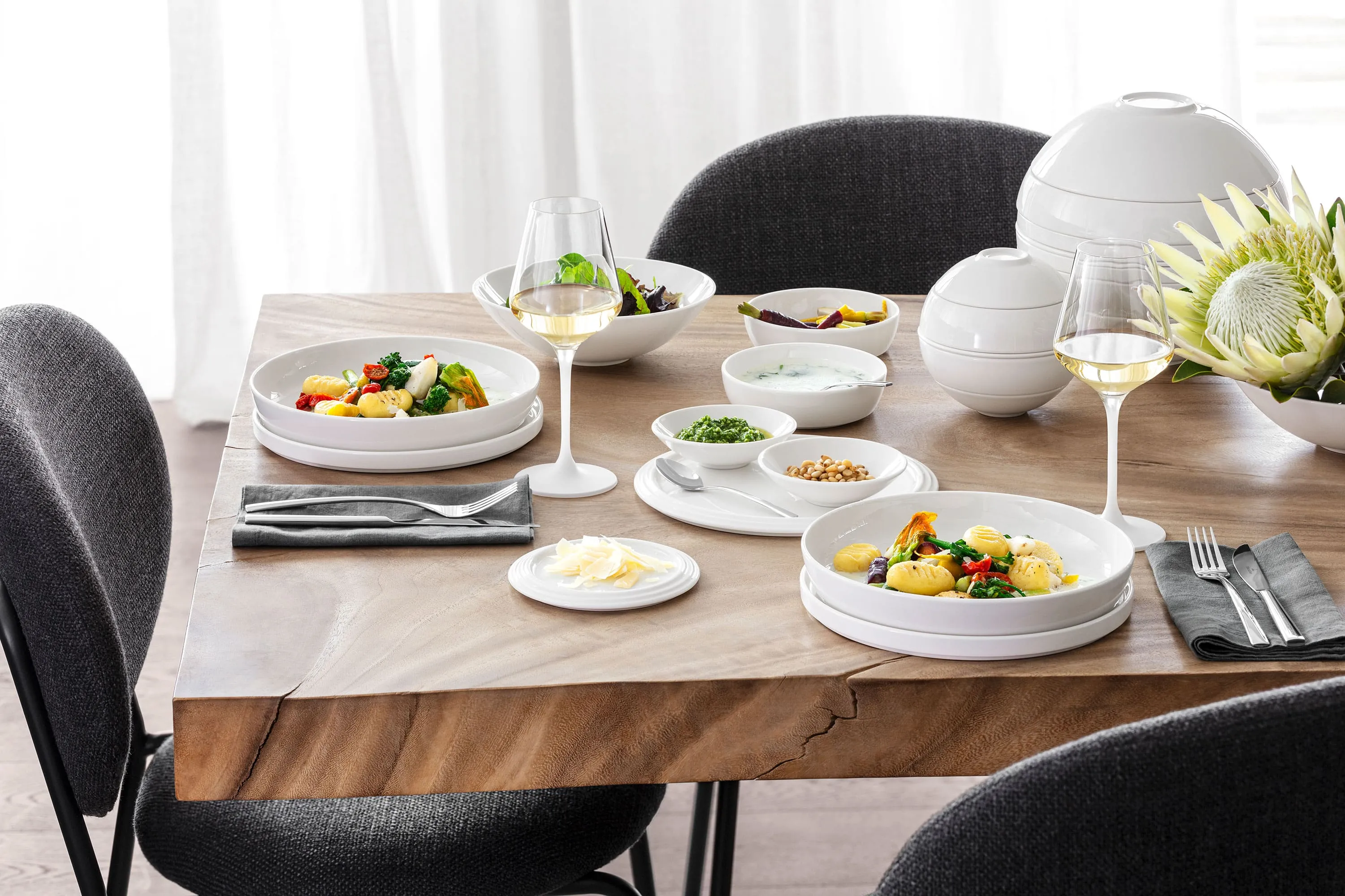 A La Boule table setting with two plates of food, two glasses of white wine, various bowls with side dishes and a flower arrangement with black and grey chairs around the table.