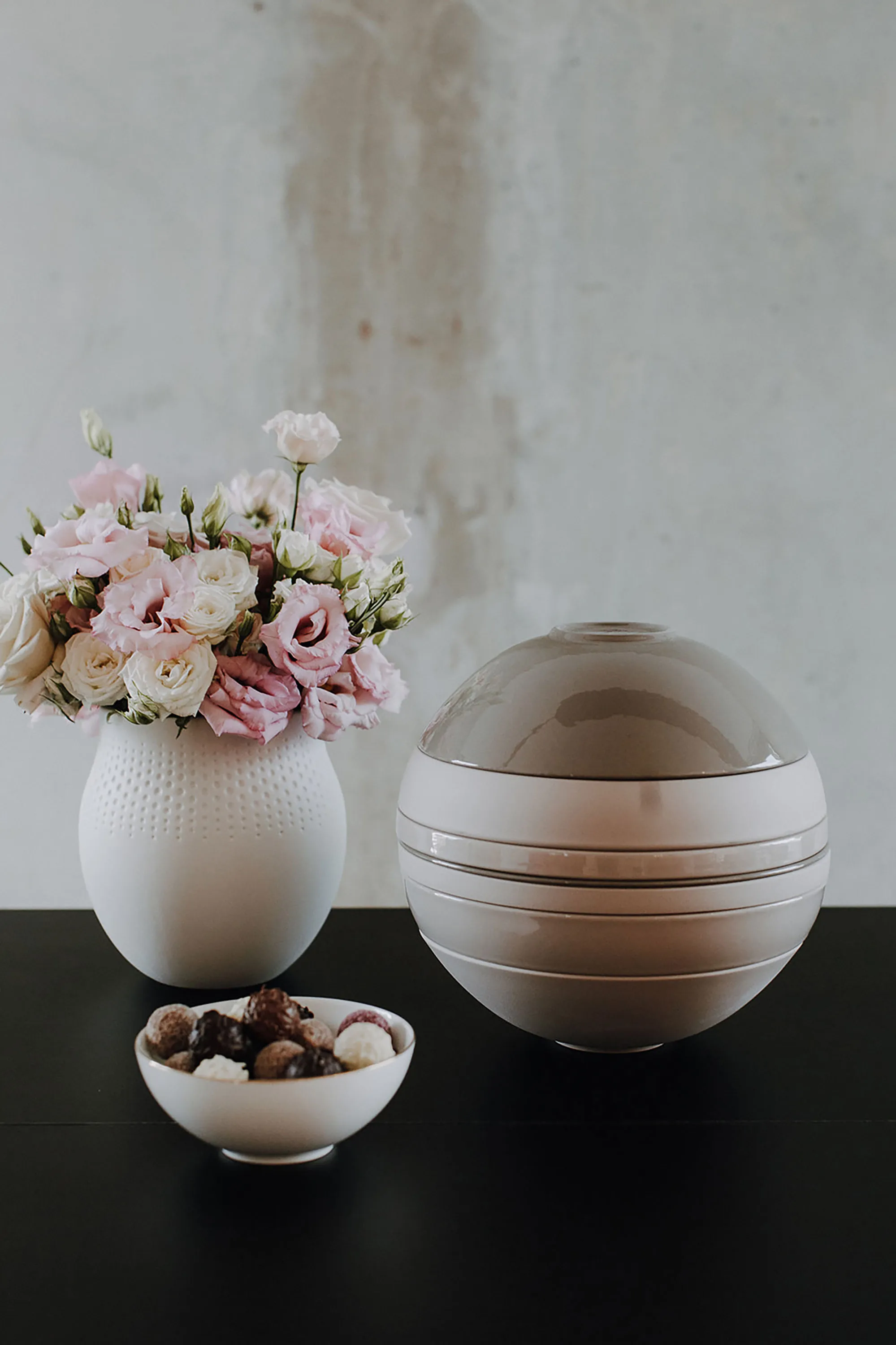 La Boule et un vase Manufacture Un vase avec des fleurs roses, un ornement rond réfléchissant et une coupe avec différentes pierres, disposées sur une surface noire.
