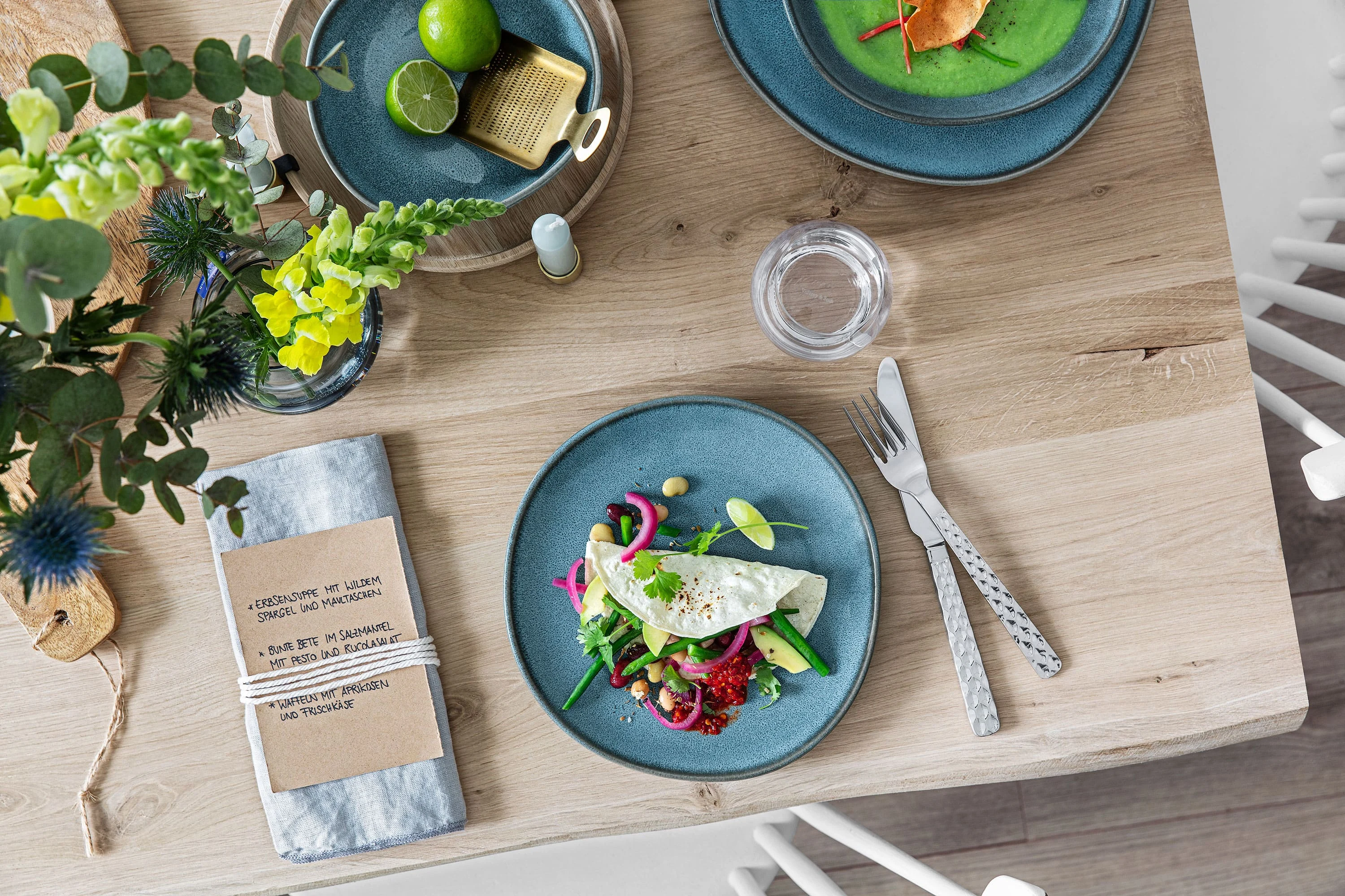 A blue plate with fresh salad, cutlery, a napkin and decorative tableware on a wooden table.