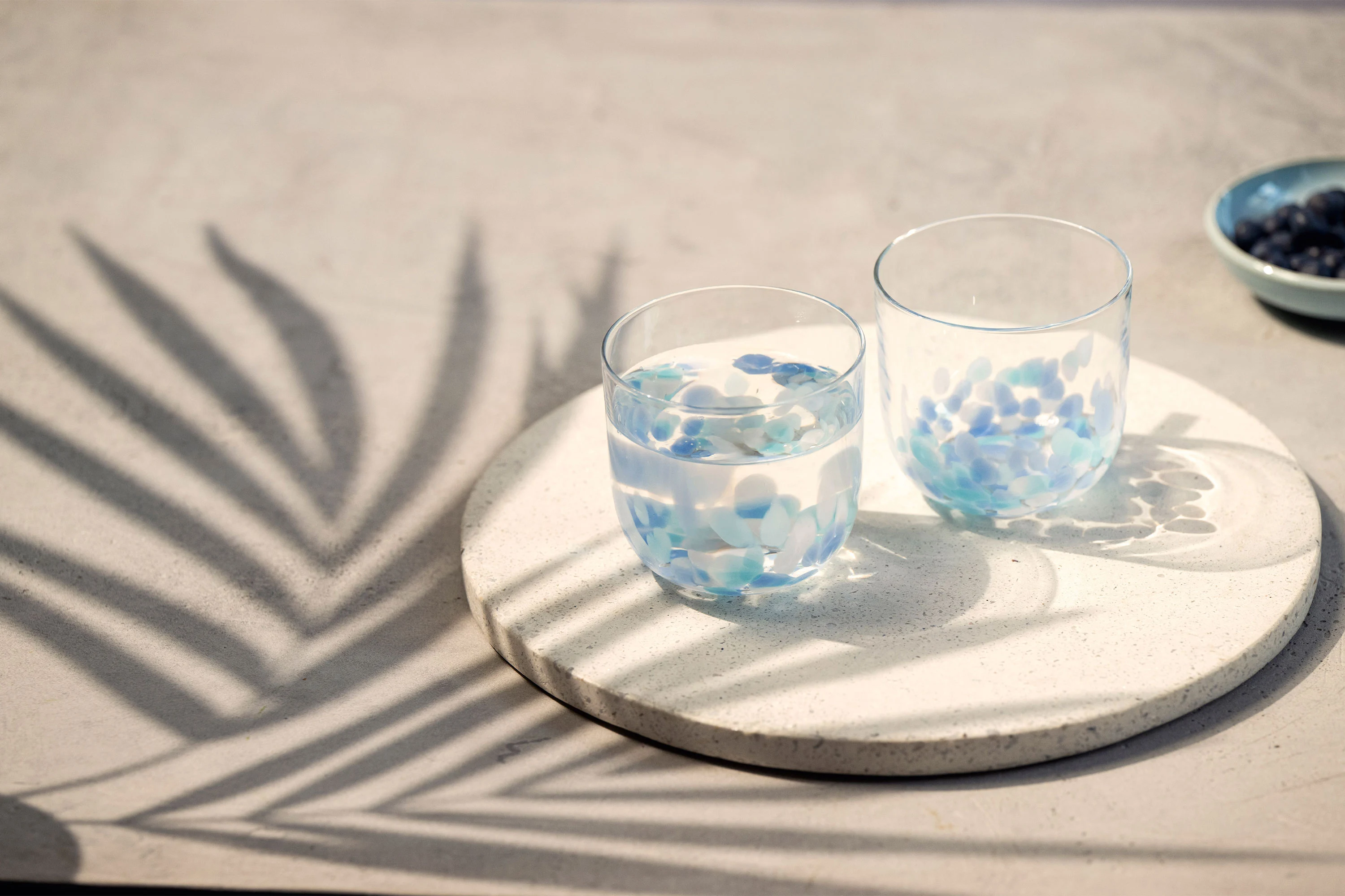 Two Like Aqua water glasses on a round tray Two Like Aqua water glasses with blue petals on a round tray, with palm leaf shadows on a light surface.