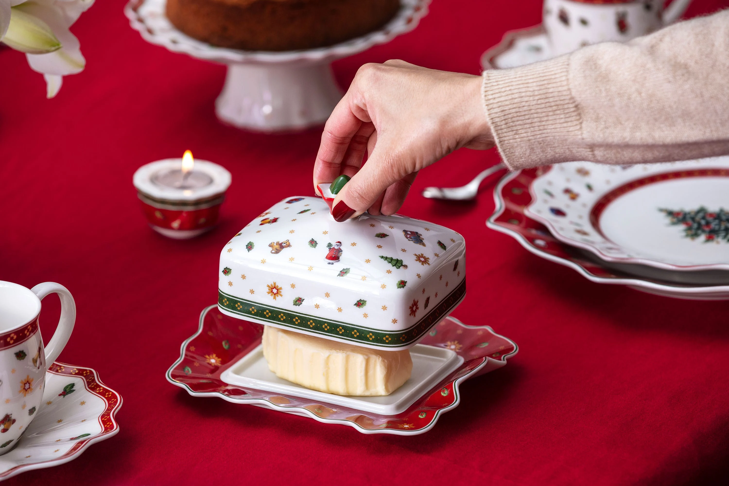 Une main soulève le couvercle d’un beurrier festif « Toy’s Delight » de Villeroy & Boch sur une nappe rouge, entourée de vaisselle ornée de motifs de Noël.