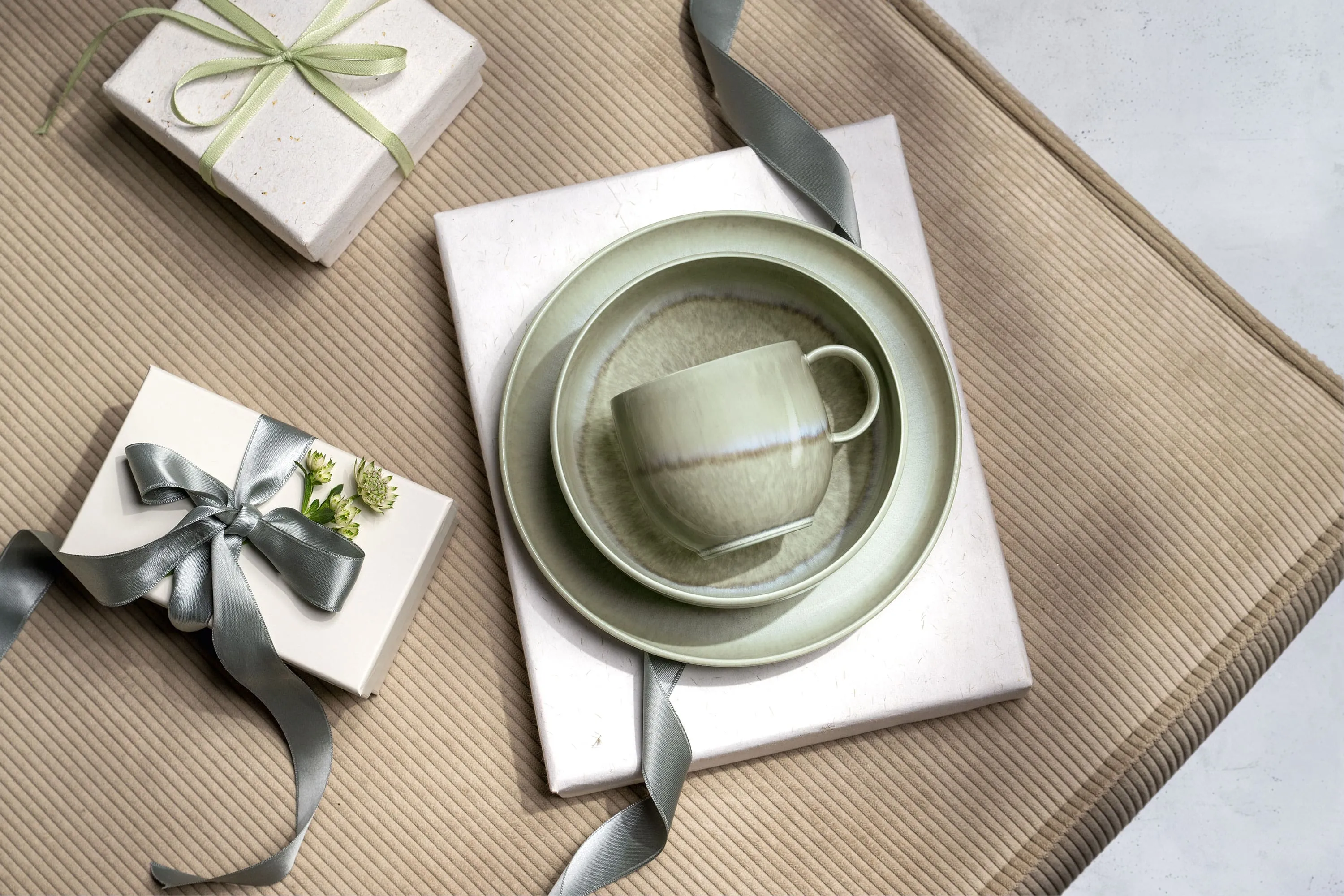 Une tasse Perlemor verte, un bol et une assiette sur un emballage cadeau Une tasse Perlemor verte, un bol et une assiette sur des cadeaux emballés avec des rubans gris, disposés sur une surface beige texturée.
