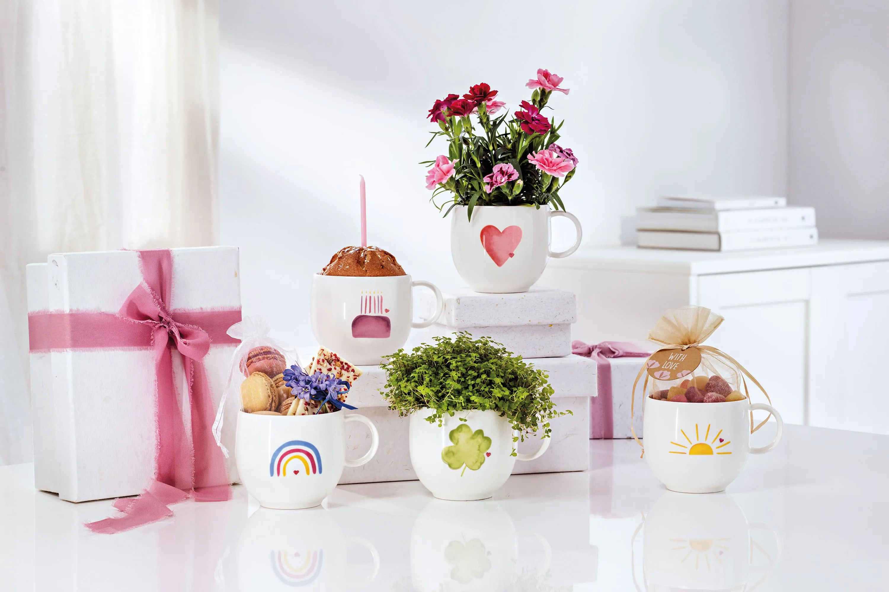 Cinq tasses blanches « With Love » de Villeroy & Boch, chacune avec un motif différent Cinq tasses blanches « With Love » de Villeroy & Boch, chacune avec un motif différent de fleurs, de sucreries et de cadeaux, sont posées sur une surface blanche. En arrière-plan, deux cadeaux emballés avec des rubans roses complètent cette scène enchanteresse.