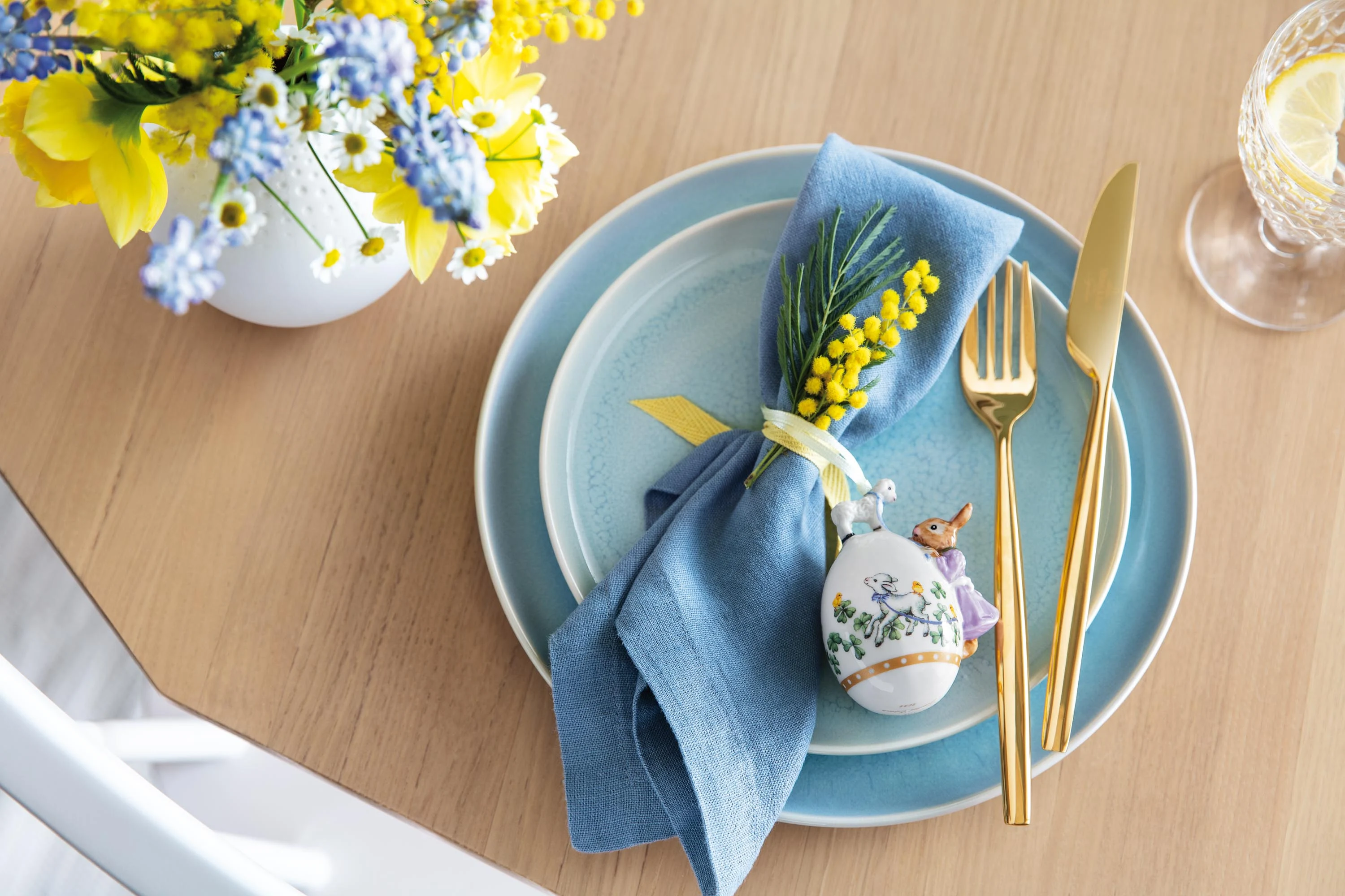 An elegantly set table with blue Crafted dinnerware and a decorative egg from the Annual Easter Edition 2022 as well as a springtime flower arrangement.