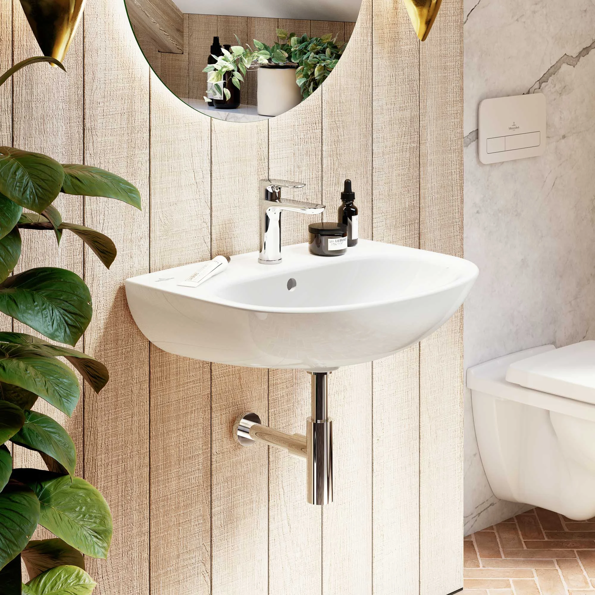 Biała umywalka O.novo z okrągłym lustrem i WC w tle Nowoczesna łazienka z białą umywalką O.novo na drewnianej ścianie, z okrągłym lustrem nad nią i różnymi przyborami toaletowymi. Po lewej stronie znajduje się roślina doniczkowa, po prawej toaleta O.novo.