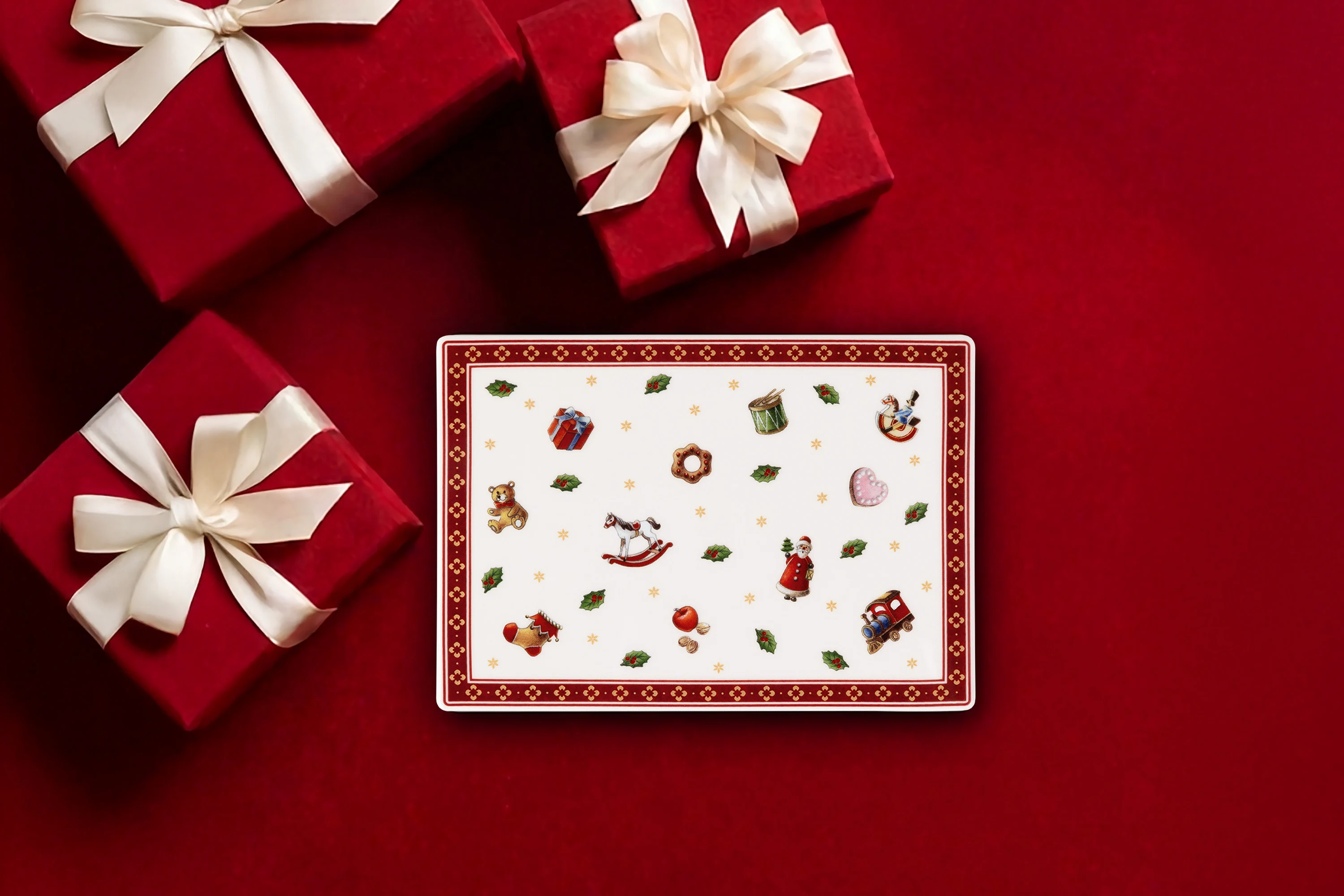 Tre scatole regalo rosse con fiocchi bianchi e un piatto rettangolare festivo su sfondo rosso.