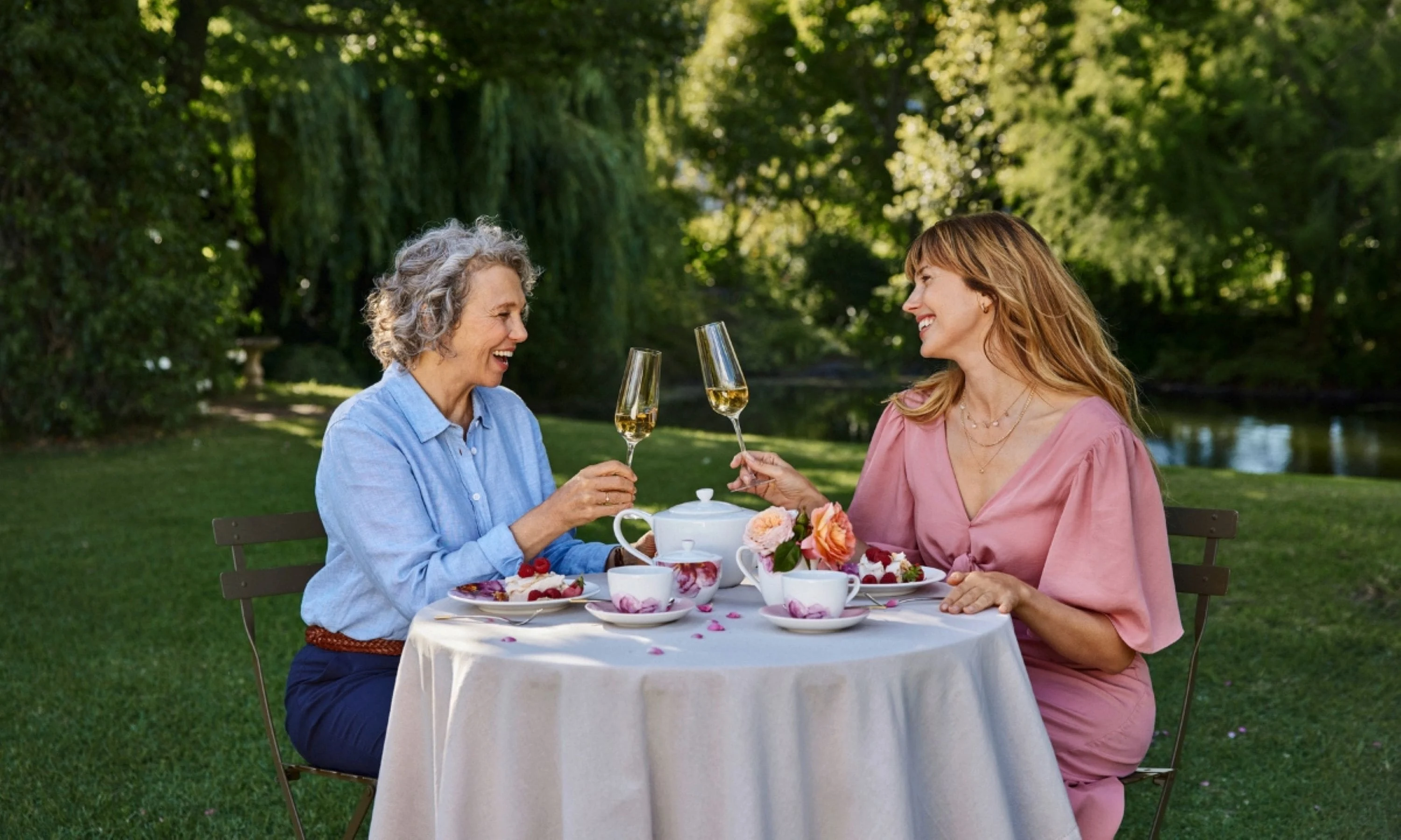 Zdjęcie Rose Garden na Dzień Matki Matka i córka siedzą na zewnątrz i wznoszą toast kieliszkami do szampana przy stole dla dwóch osób z czajnikiem i filiżankami we wzór Rose Garden