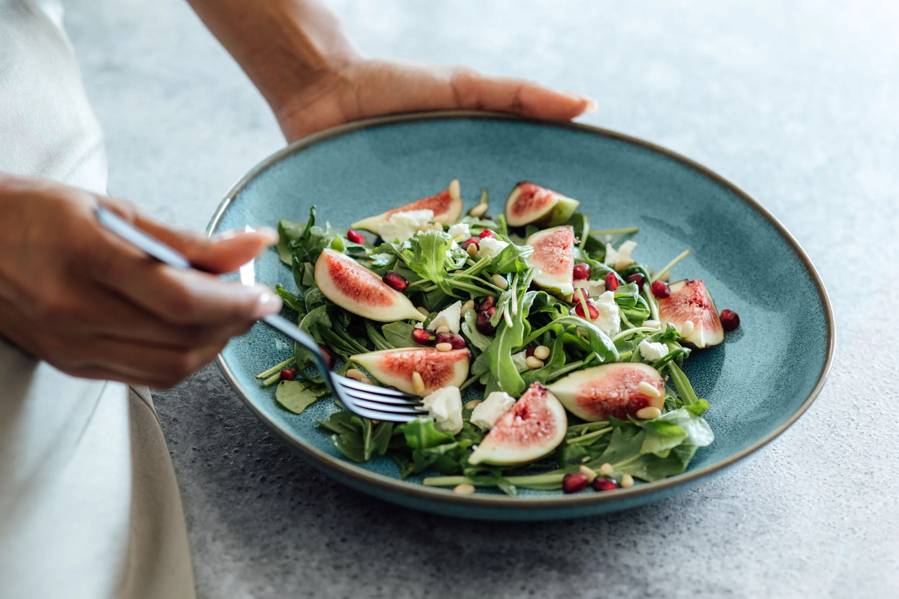 Someone is holding a plate of salad—figs, arugula, pine nuts, pomegranate seeds—and eating with a fork.