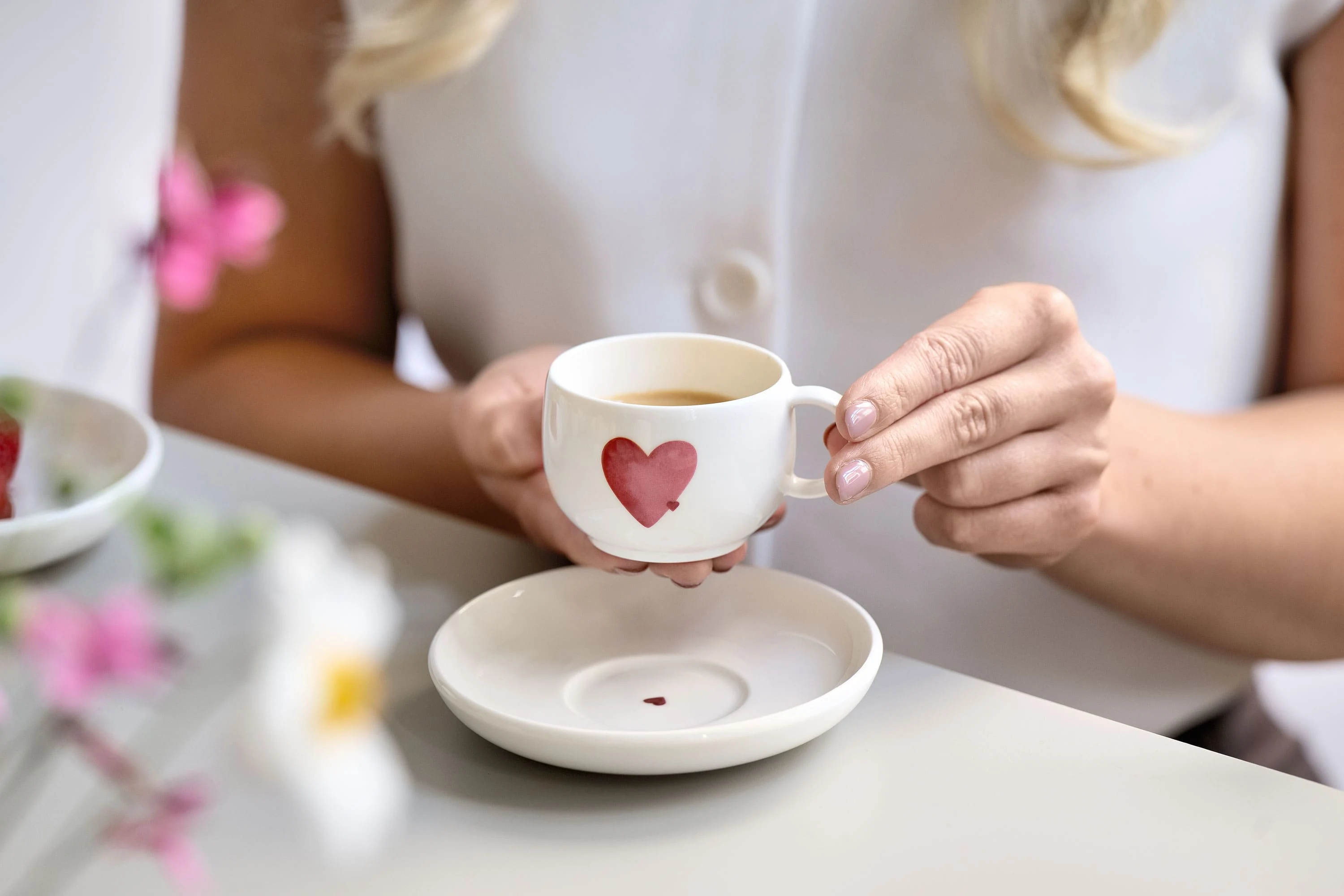 Eine Person hält eine weiße Kaffeetasse mit einem roten Herzmotiv über einer passenden Untertasse mit rotem Herzmotiv auf einem Tisch.