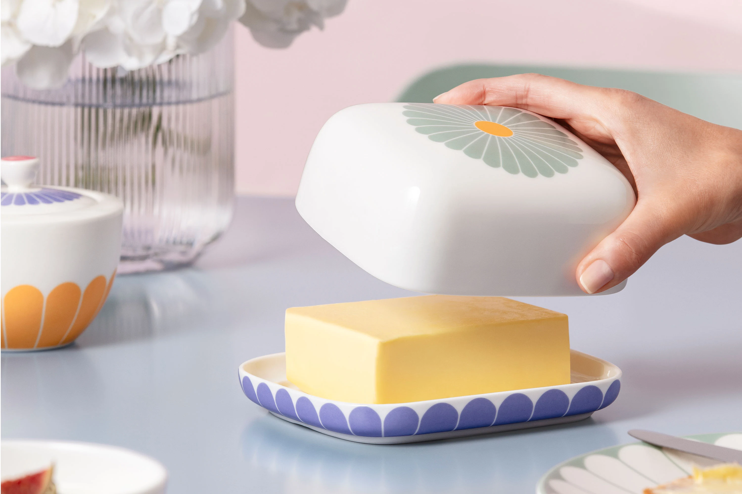 Une main soulève le couvercle du beurrier en céramique Fleur, dans lequel repose un morceau de beurre sur une table.
