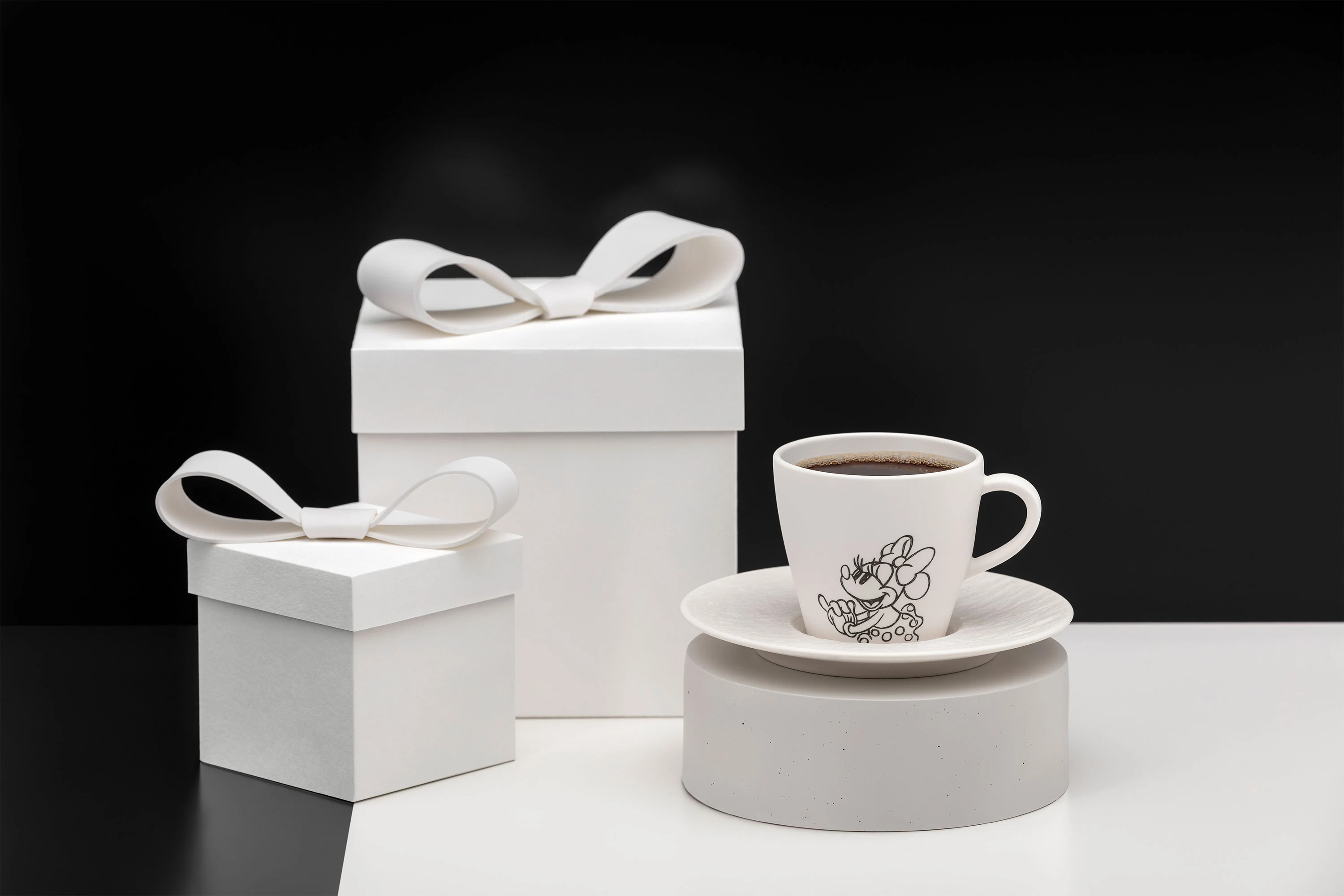 Zwei weiße Geschenkboxen mit Schleifenband und eine Manufacture Mickey Kaffeetasse auf einer Untertasse vor einem schwarz-weißen Hintergrund.
