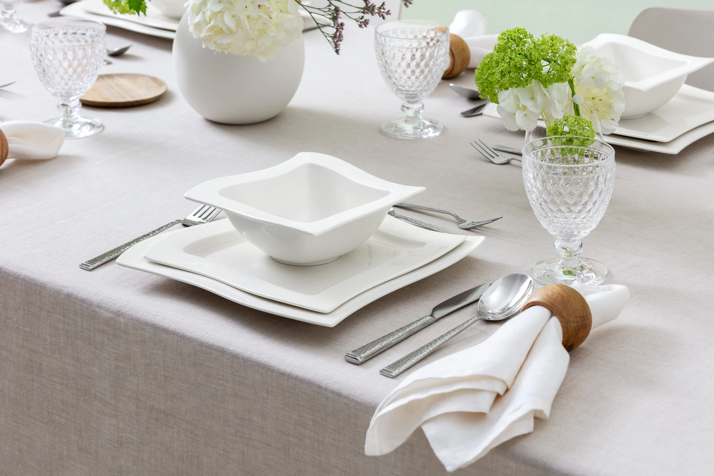 Fine table setting with white square NewWave plates, glasses, and a napkin with a wooden ring. Floral arrangement with white and green flowers on a light grey tablecloth.