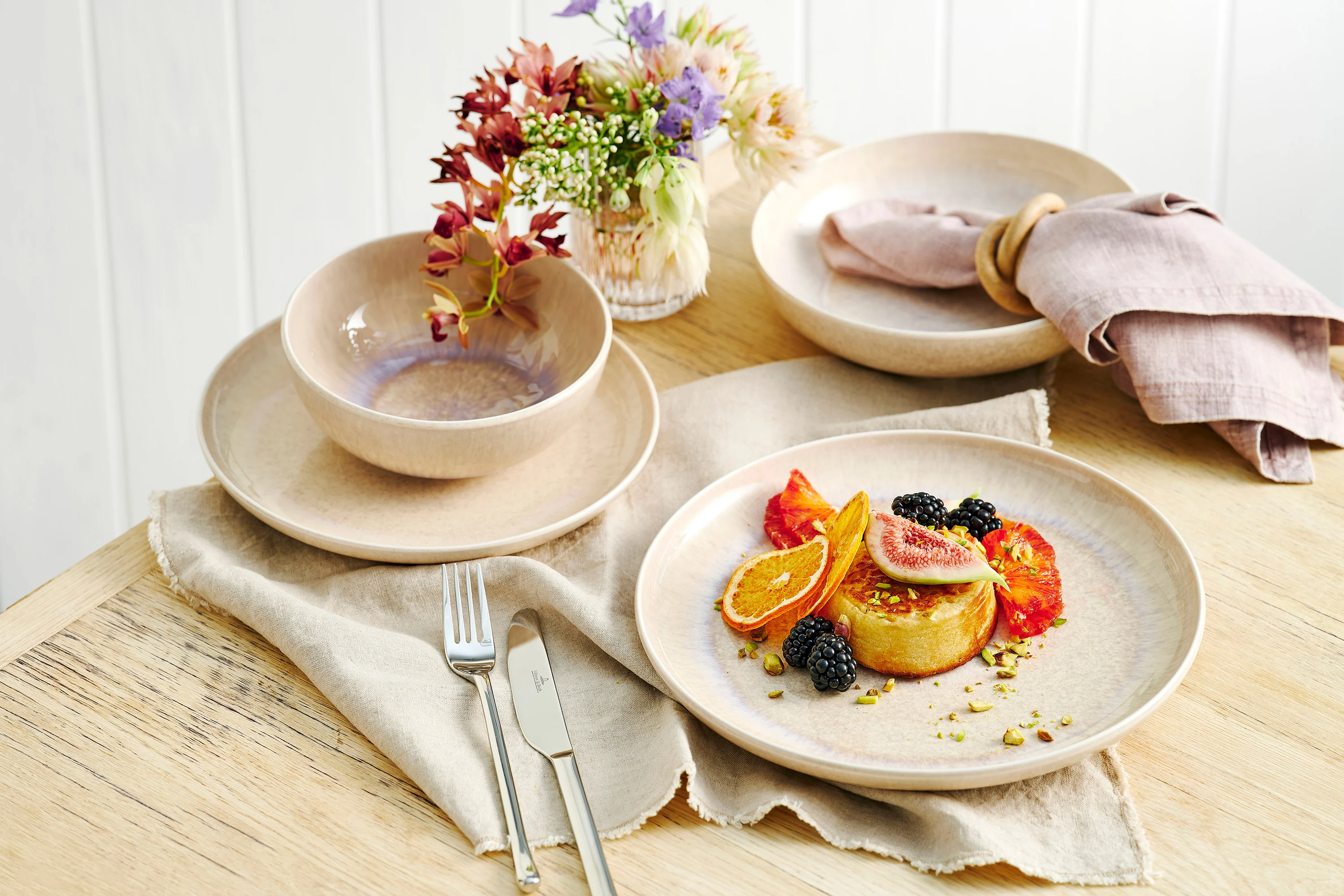 aus-perlemorsand-indoor-food-nopeople-dinnerset7069 The Perlemor Sand dinner set for one, featuring a plate filled with a dessert topped with fresh fruits, rests on a brown linen tablecloth atop a rustic wooden table, paired with silver cutlery.