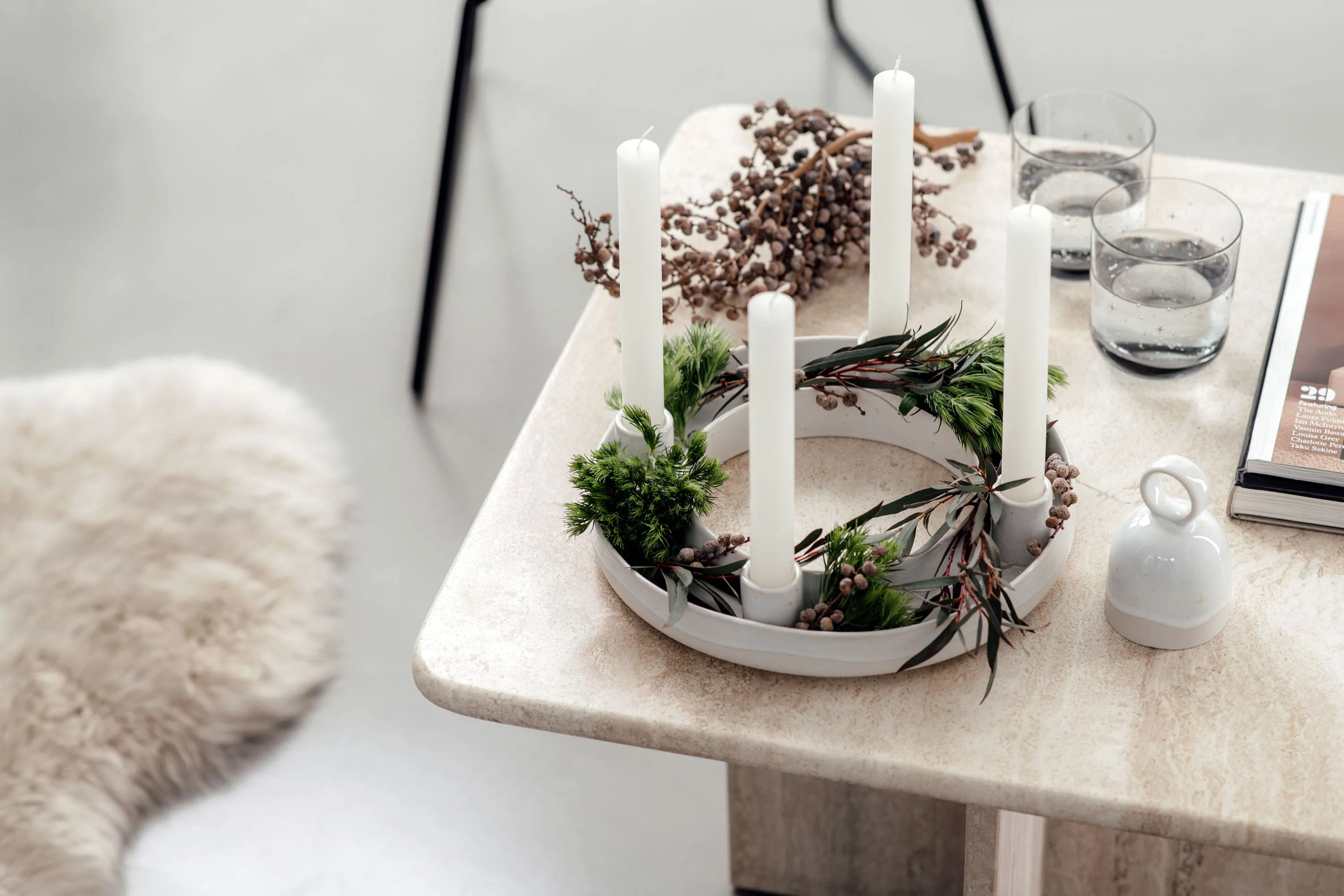 Une table beige avec une couronne de l’Avent Winter Glow au centre, entourée de verres Winter Glow et d’un magazine.