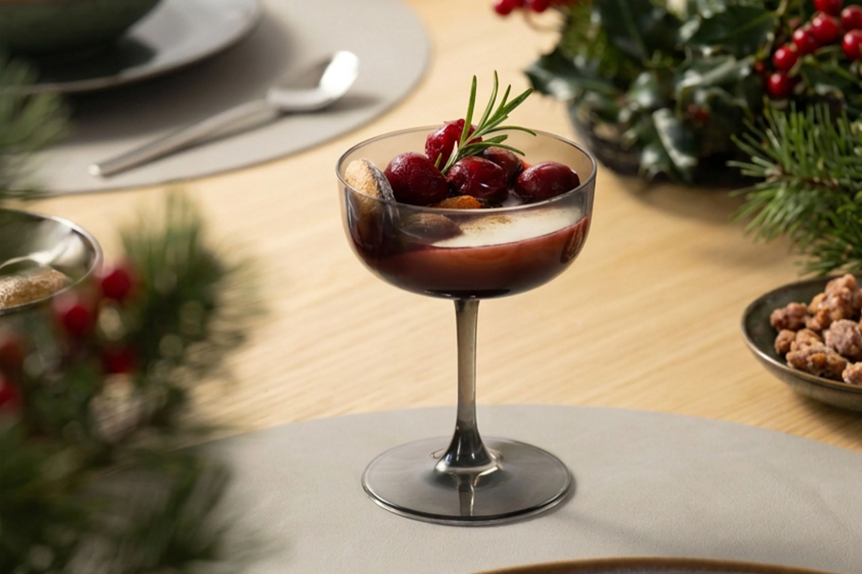 Primer plano de una copa de champán Like Glass de Villeroy & Boch en color Smoke, utilizada como copa de postre para una crema navideña en capas con cerezas rojas y una ramita de romero, sobre una mesa de comedor moderna.