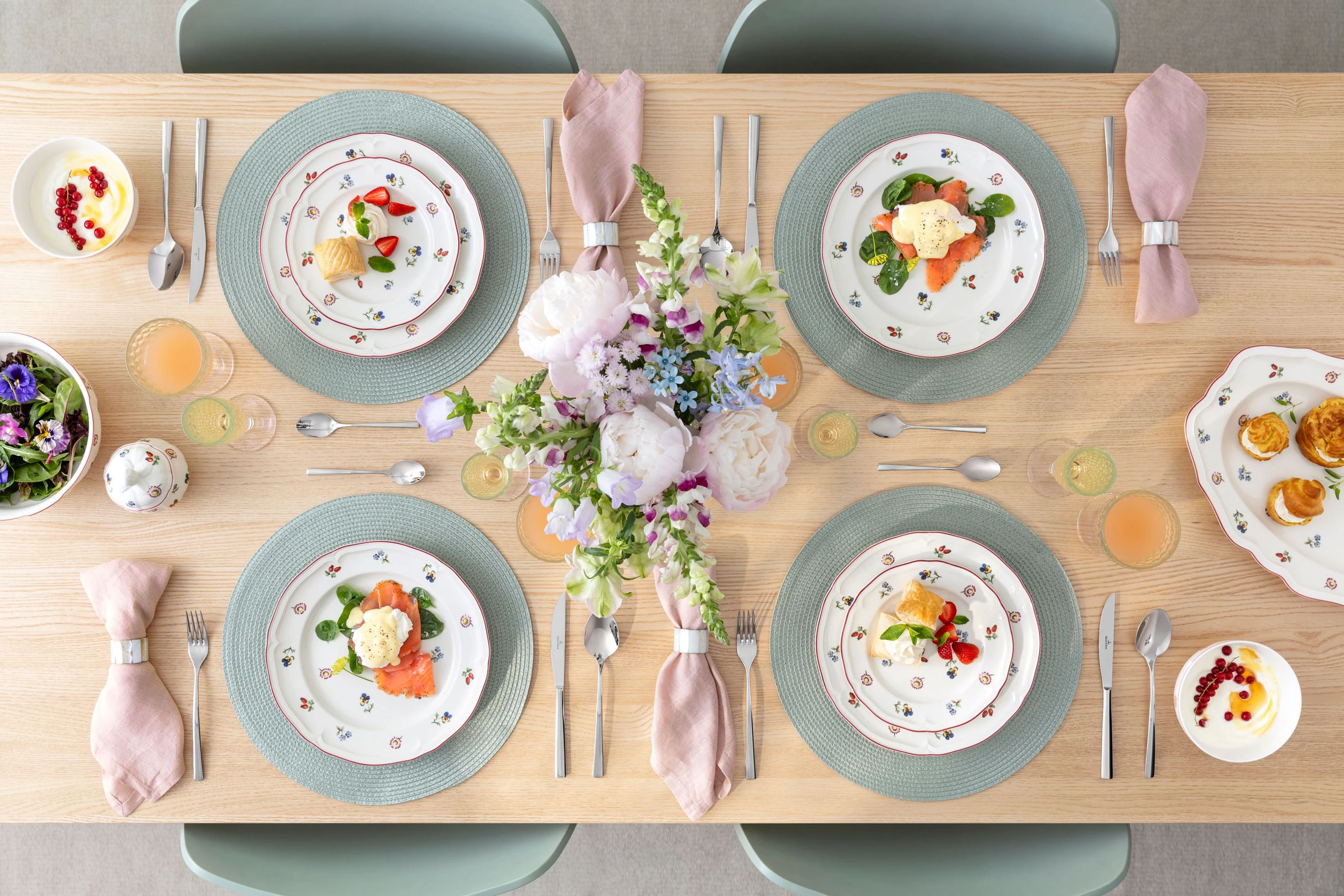 Ein gedeckter Esstisch für vier Personen mit Petite Fleur Blumentellern, rosa Servietten, Getränken und einem Blumenschmuck.