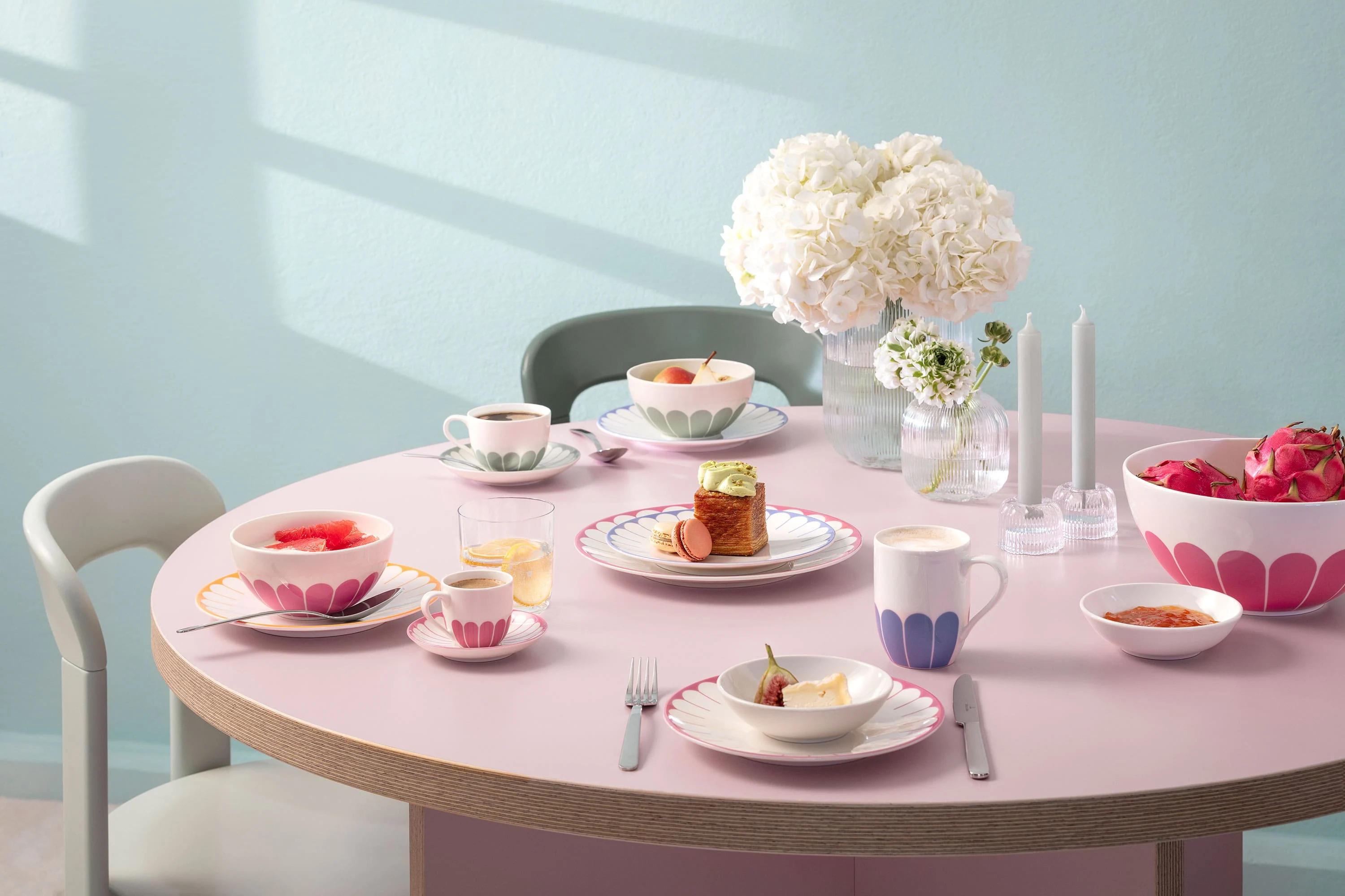Une table ronde avec des assiettes, des tasses et des bols remplis de nourriture, décorés de motifs roses. Au centre se trouvent un vase avec des fleurs blanches et deux bougies non éclairées. Des chaises entourent la table.
