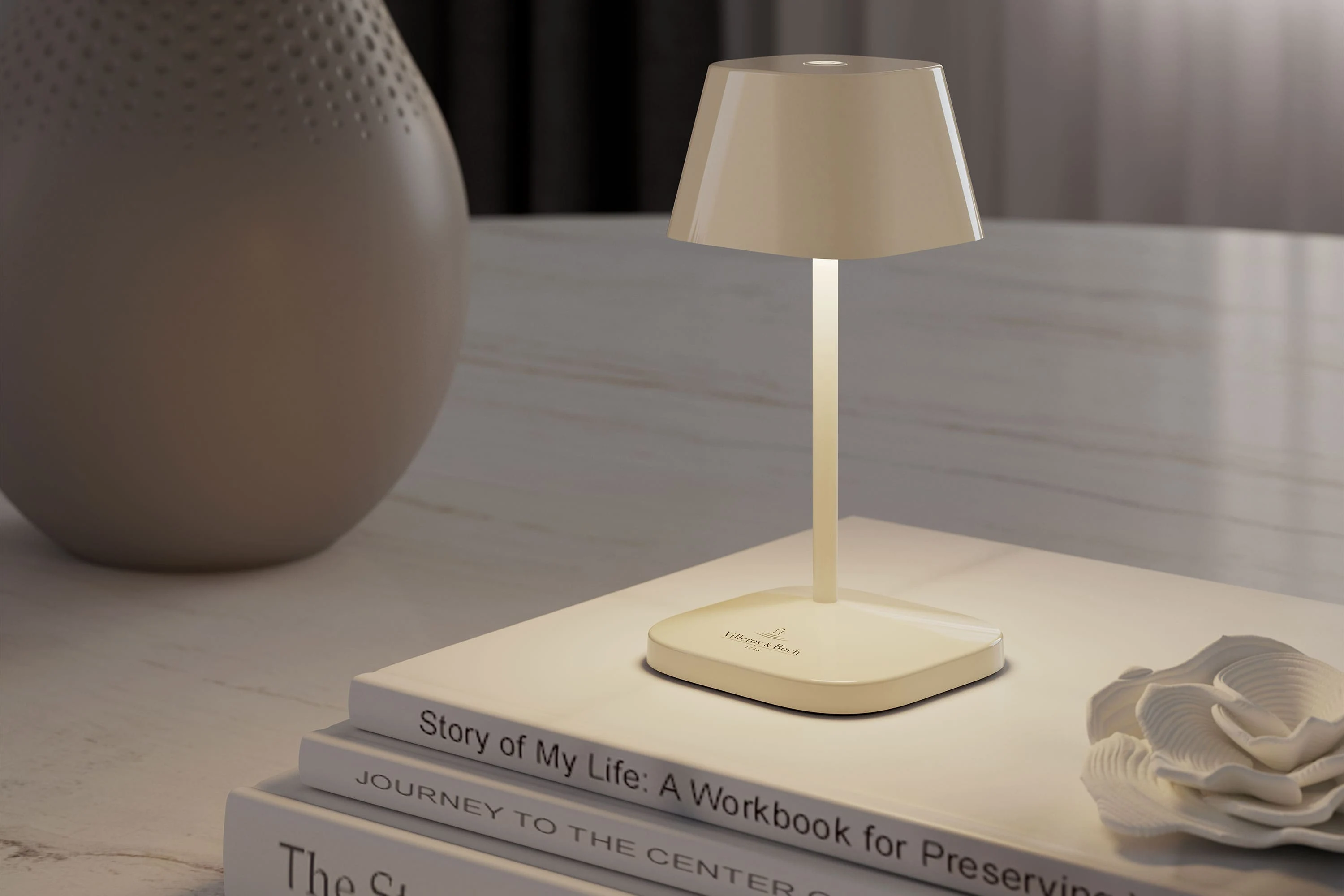  A small, modern table lamp on stacked books next to a decorative vase and a white flower on a bright surface. 
