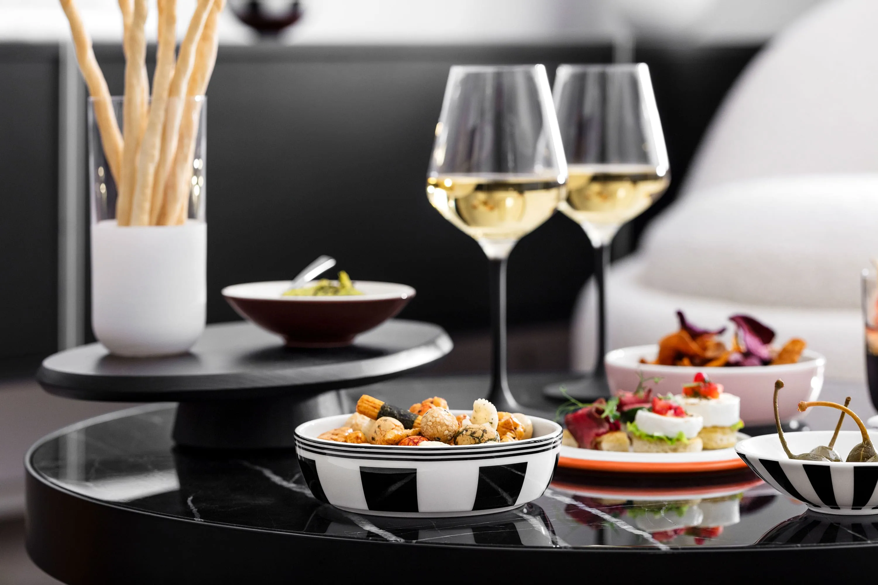 A black table with La petite Boule individual bowls filled with nibbles and Manufacture white wine glasses.