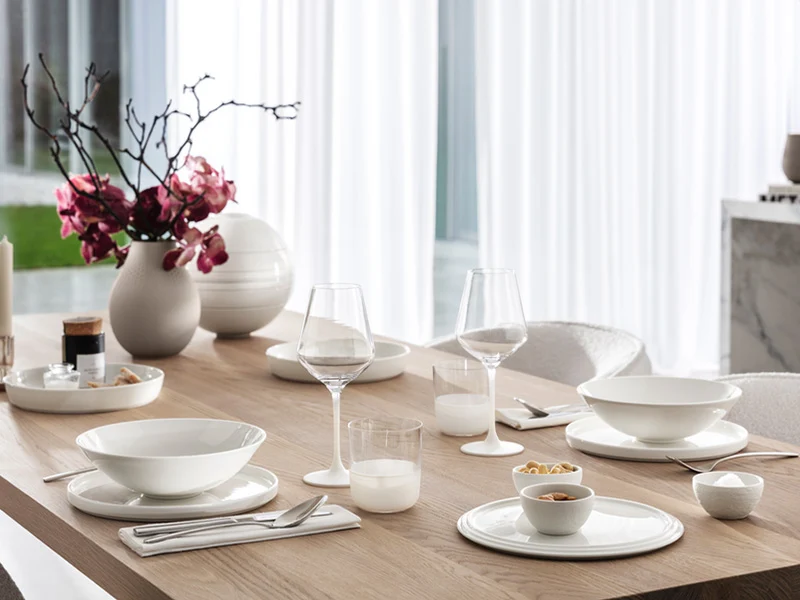 Vaisselle La Boule et verres Manufacture Une table de salle à manger moderne avec de la vaisselle blanche, des verres et un vase avec des fleurs roses sur une surface en bois.