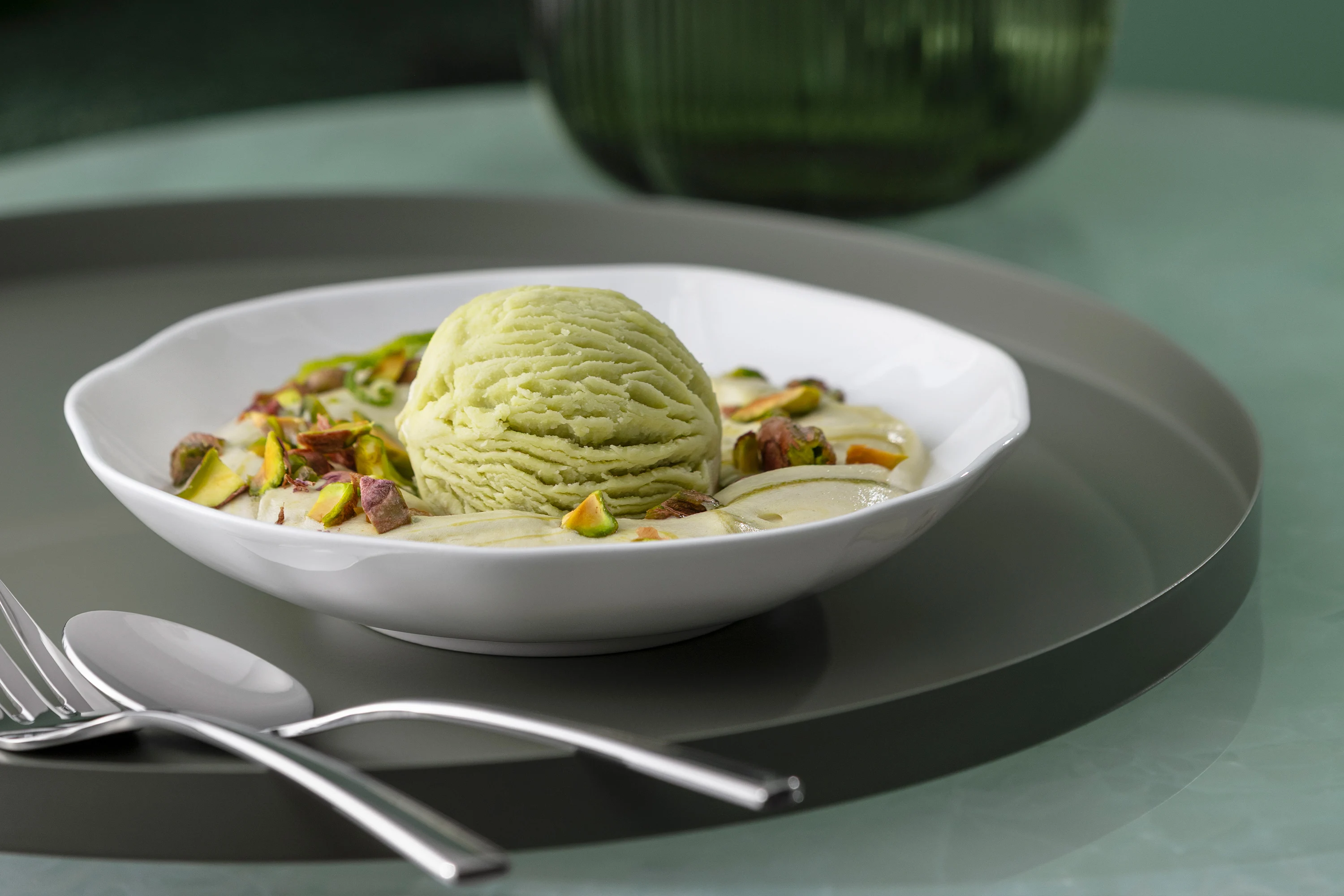 A Pura dessert bowl with pistachio ice cream A Pura dessert bowl with pistachio ice cream, chopped pistachios and cream on a white plate with cutlery beside it.