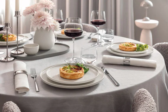 A Villeroy & Boch Afina table set for four with quiche plates, wine glasses, napkins and a vase with pink flowers.